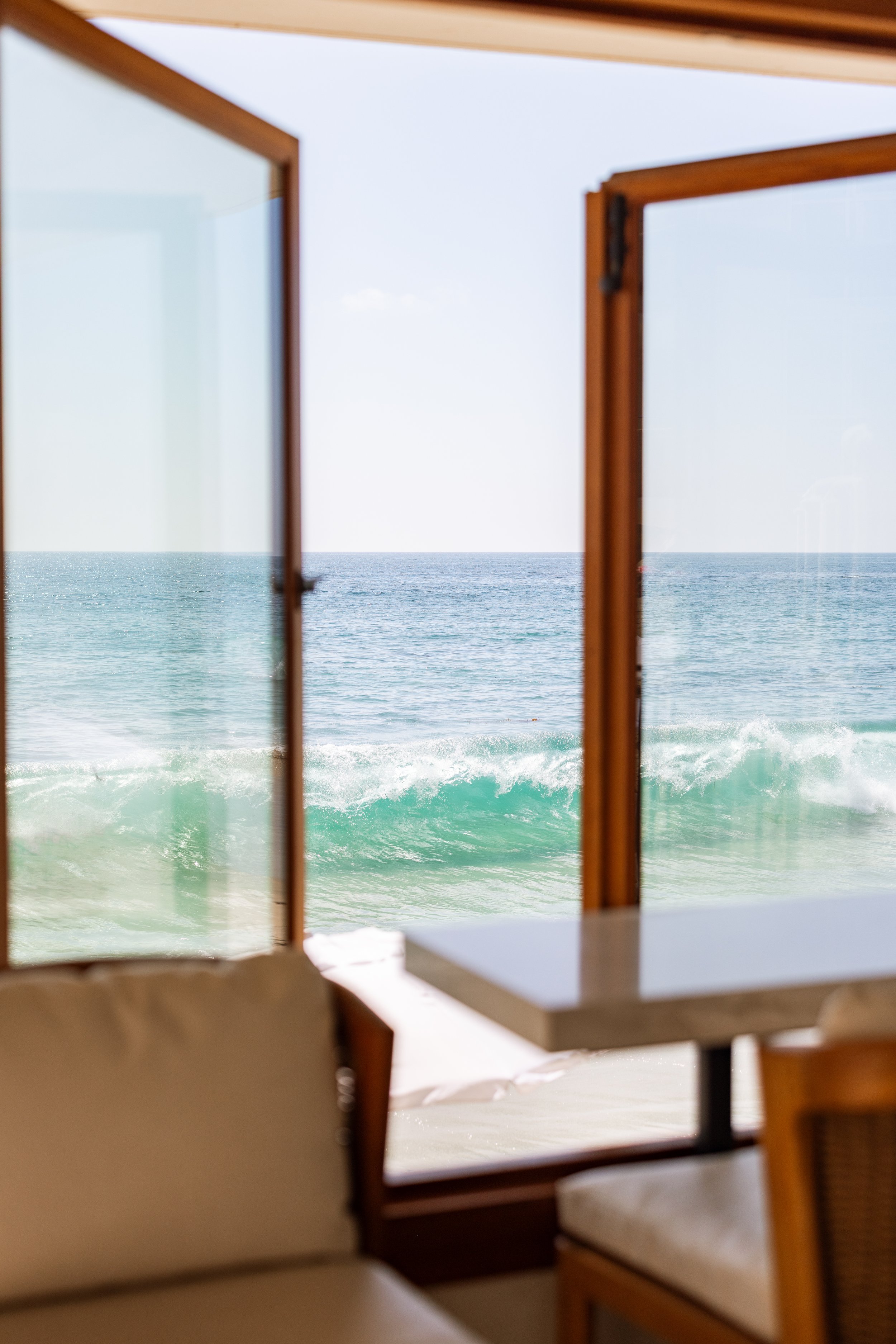 View of the ocean through open wooden window frames from inside a room, with a table and seating area in the foreground. Photo by Summerside Creative A Hotel Social Media & Creative Agency Serving Hospitality Brands