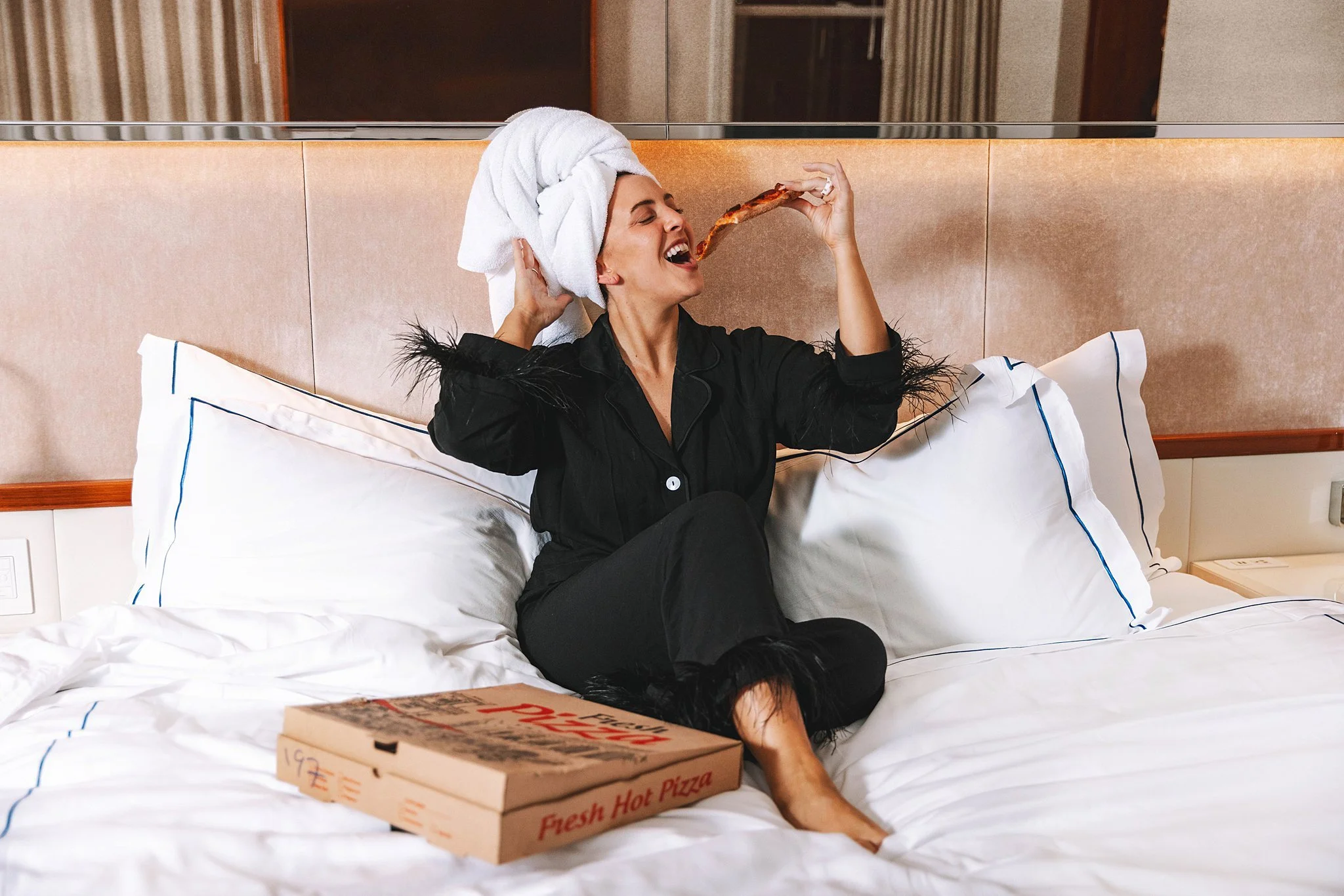 Woman sitting on a bed with a towel in her hair, wearing black pajamas, holding pizza near her open mouth, with a pizza box on the bed. Photo by Summerside Creative A Hotel Social Media & Creative Agency Serving Hospitality Brands
