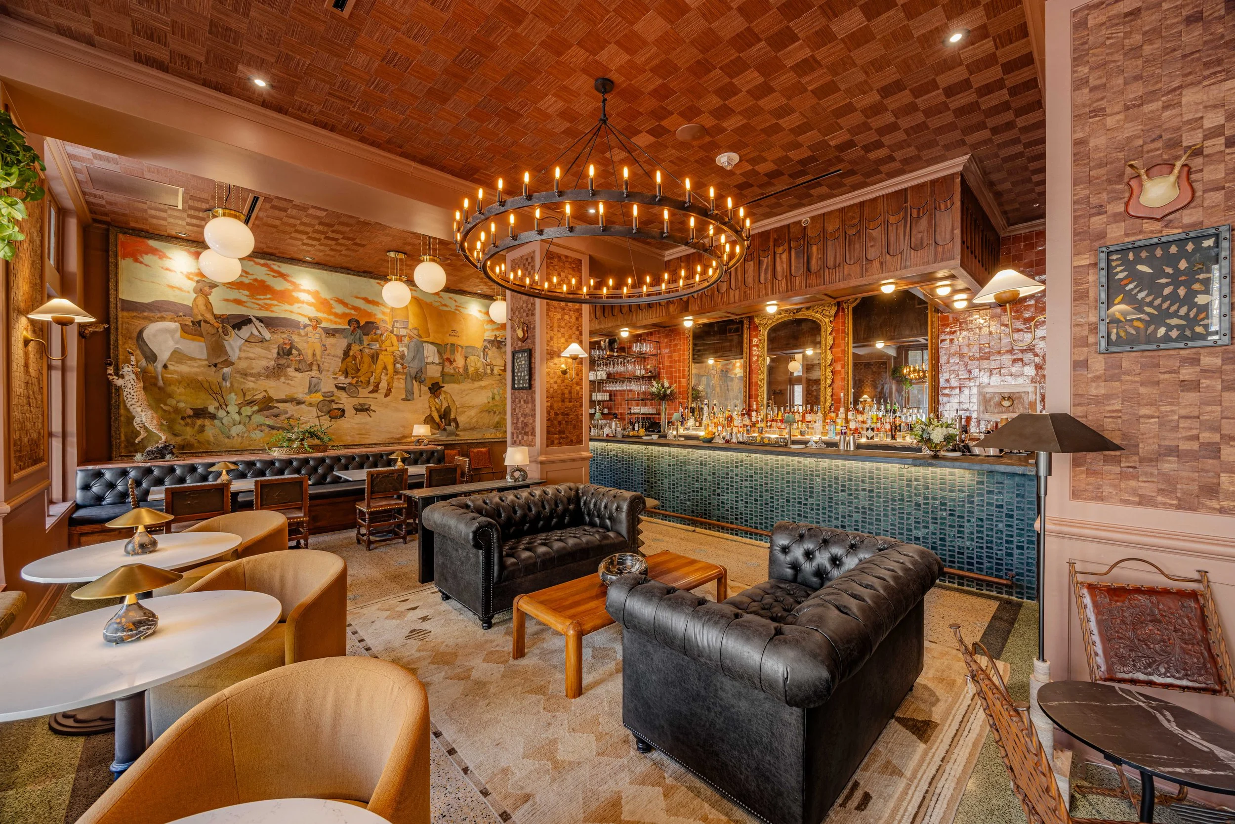 Interior of a cozy bar with leather seating, a large chandelier, a bar counter with bottles, vintage artwork, and warm lighting. Photo by Summerside Creative A Hotel Social Media & Creative Agency Serving Hospitality Brands 