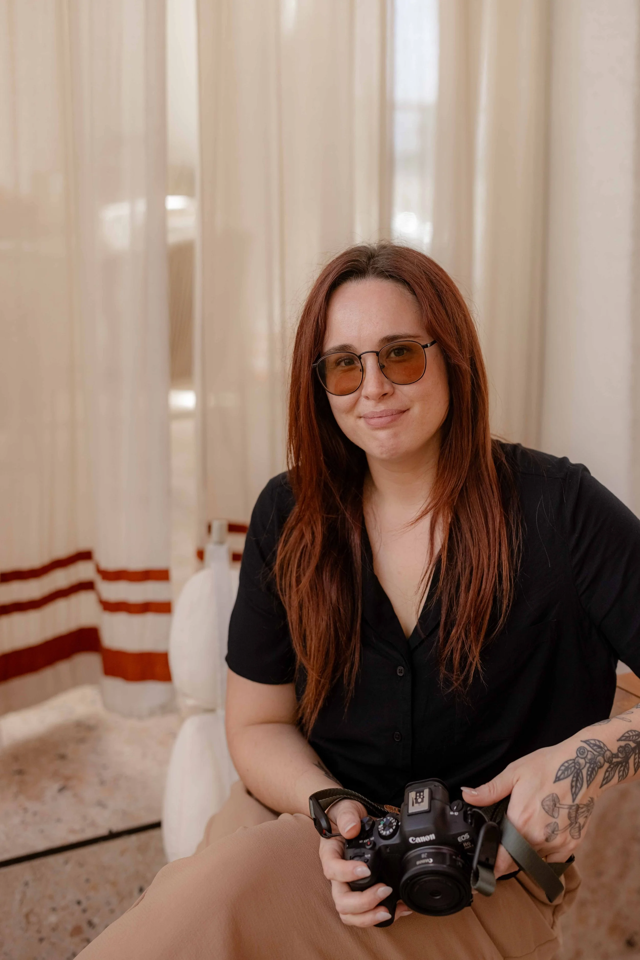 A woman with long red hair and glasses, wearing a black shirt and beige pants, sitting indoors with a camera in her hand, smiling slightly at the camera.