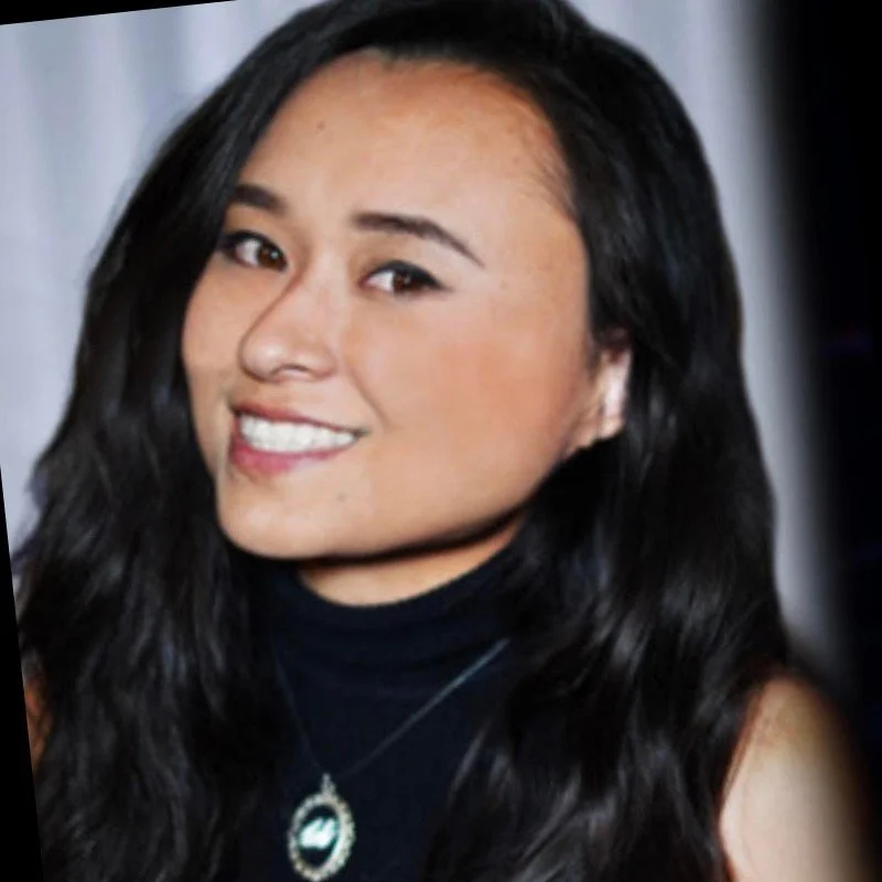A young woman with long black hair smiling at the camera, wearing a black top and a necklace with a circular pendant.