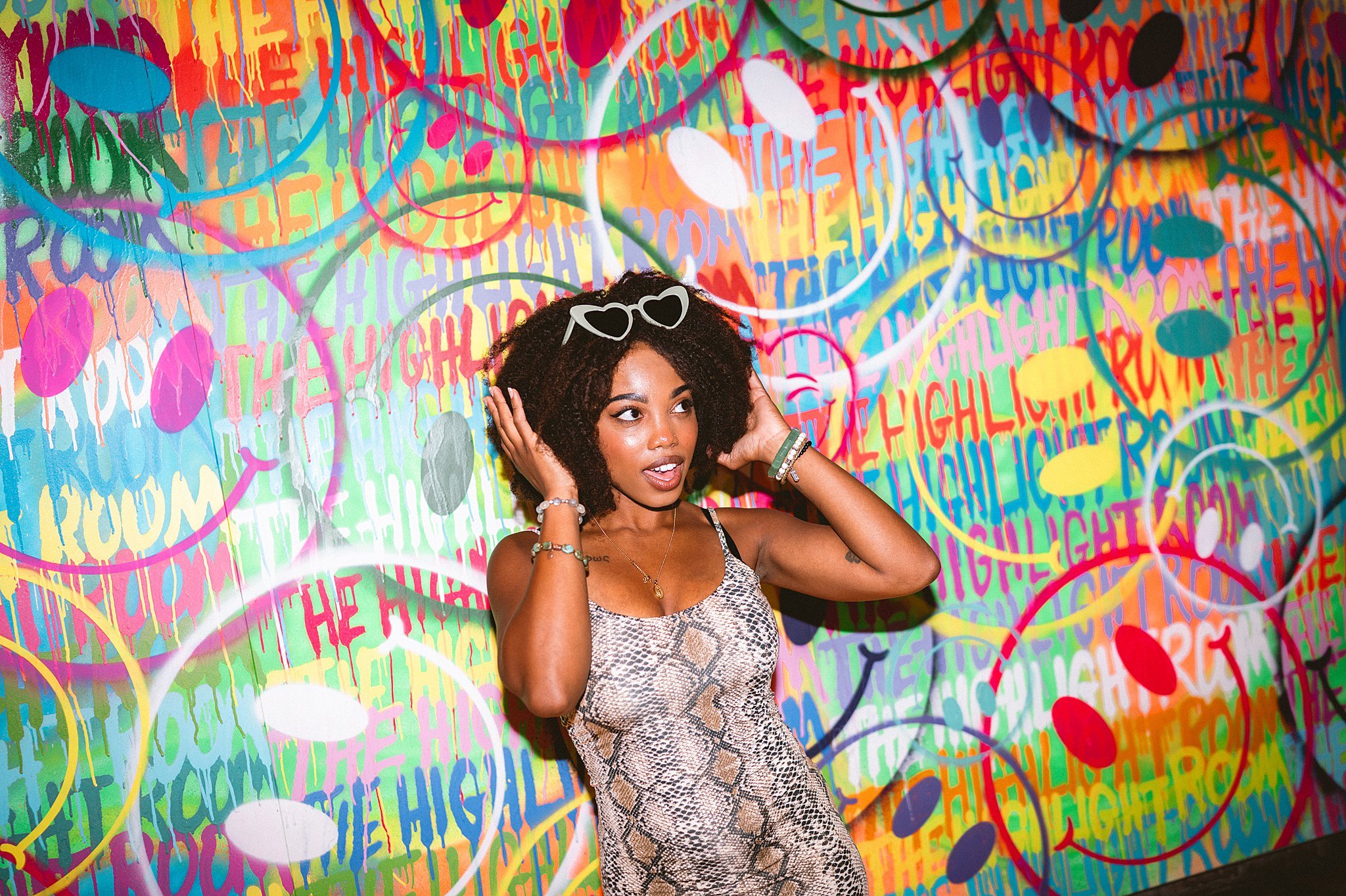 A young woman with curly hair wearing sunglasses on her head, a snake print dress, and jewelry, standing in front of a colorful graffiti wall. Photo by Summerside Creative A Hotel Social Media & Creative Agency Serving Hospitality Brands