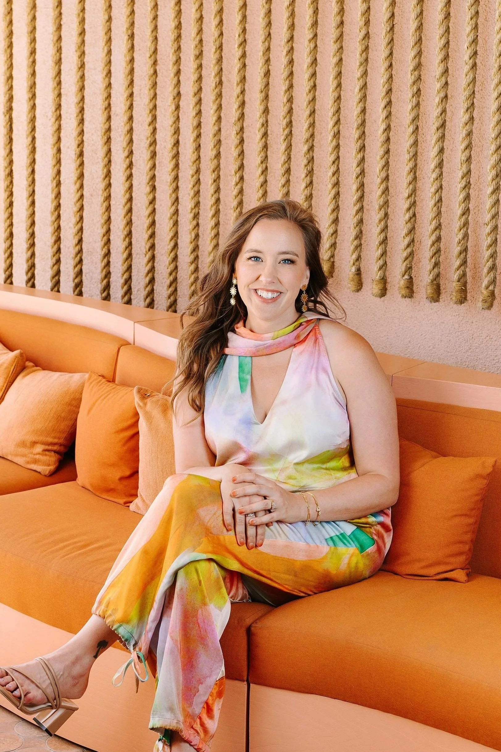 A woman with long wavy hair wearing an orange satin blouse and dark jeans standing indoors against a brick wall with a bicycle mounted above her. Photo by Summerside Creative A Hotel Social Media & Creative Agency Serving Hospitality Brands