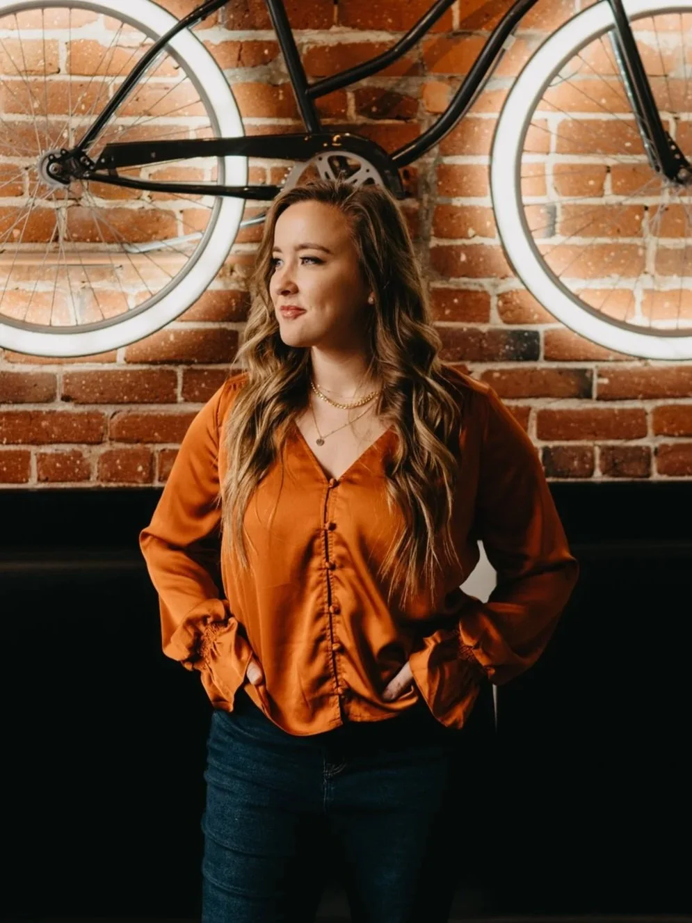 A woman with long wavy hair, wearing an orange satin blouse and dark jeans, stands with her hands on her hips in front of a brick wall with a bicycle mounted above her, illuminated by neon lights.
