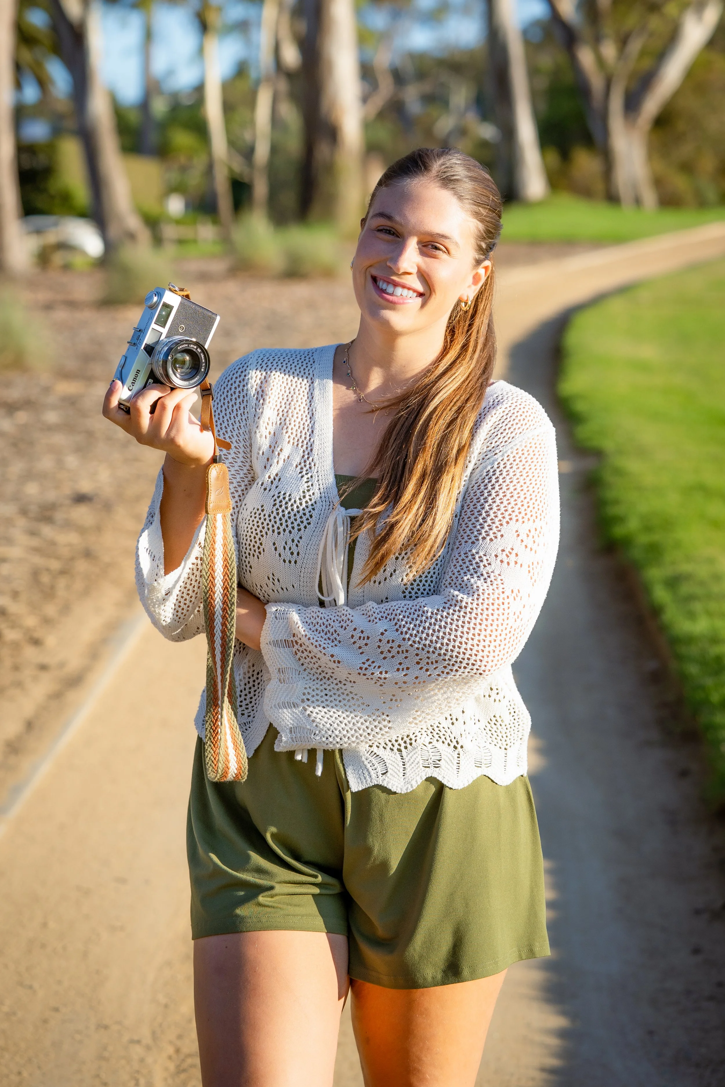 A young woman standing outdoors on a sunny day, holding a vintage camera and smiling.