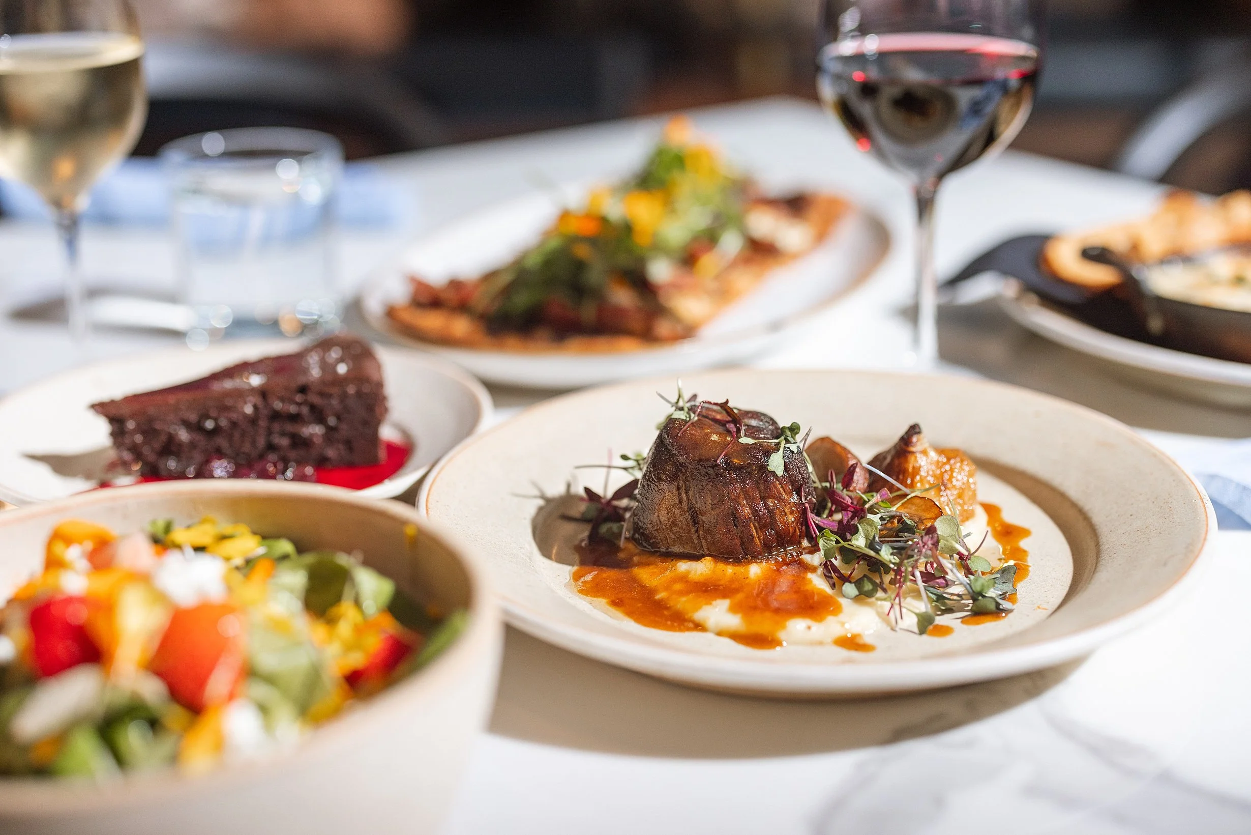 A table with various dishes including a piece of chocolate cake, a plate of braised meat with sauce and microgreens. Photo by Summerside Creative A Hotel Social Media & Creative Agency Serving Hospitality Brands
