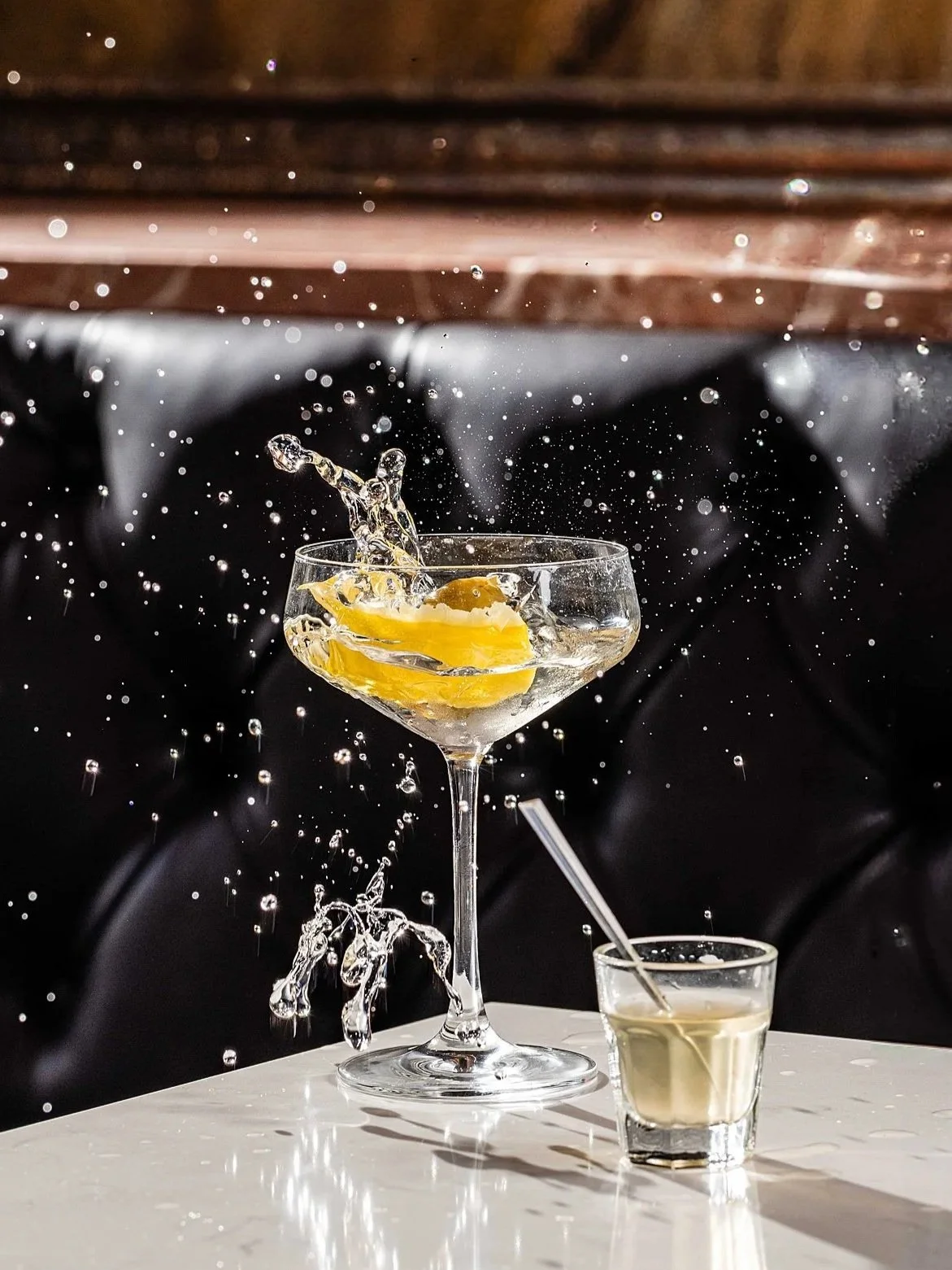 A splash of water and lemon slice in a cocktail glass on a white table, with a smaller shot glass and straw beside it, against a dark background. Photo by Summerside Creative A Hotel Social Media & Creative Agency Serving Hospitality Brands 
