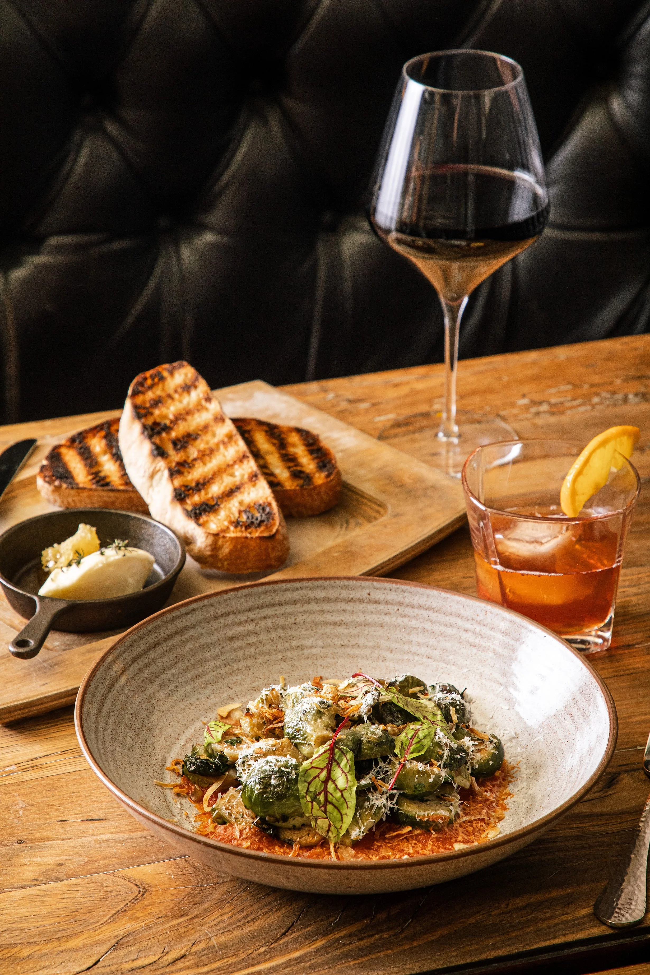 A wooden table with a bowl of pasta topped with greens, a glass of red wine, a glass of cocktail with lemon, and a wooden board. Photo by Summerside Creative A Hotel Social Media & Creative Agency Serving Hospitality Brands 