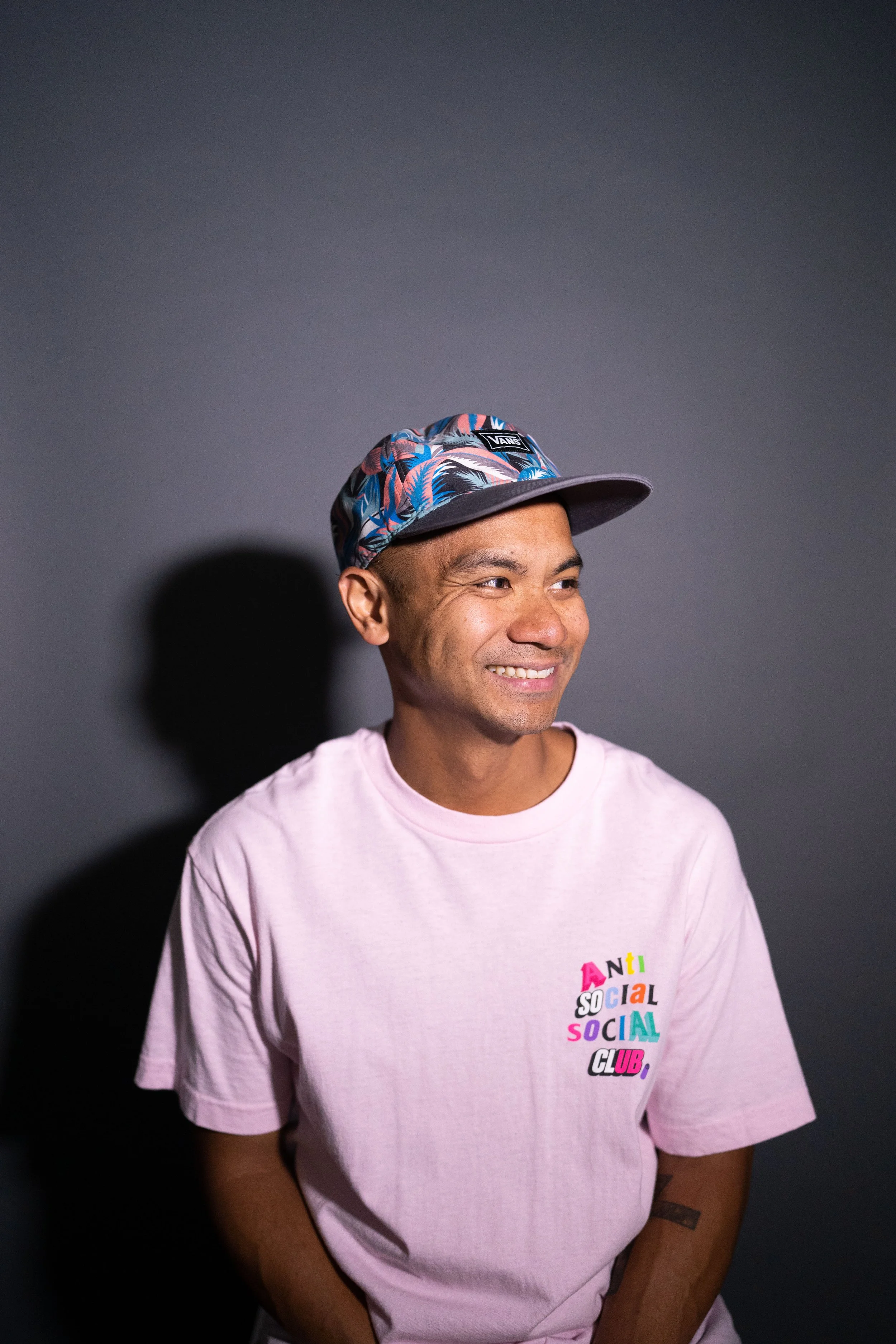 A young man with a cap and pink t-shirt smiling against a dark background.