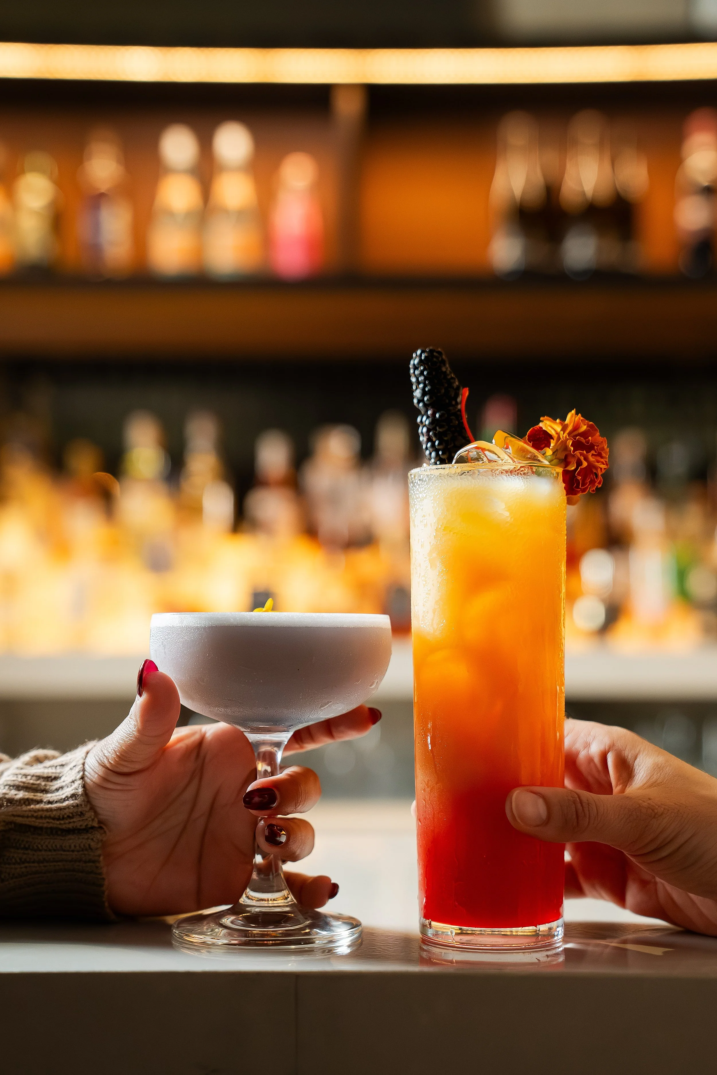 Two cocktails on a bar, one in a coupe glass and one in a tall highball glass, with blurred liquor bottles in the background. Photo by Summerside Creative A Hotel Social Media & Creative Agency Serving Hospitality Brands