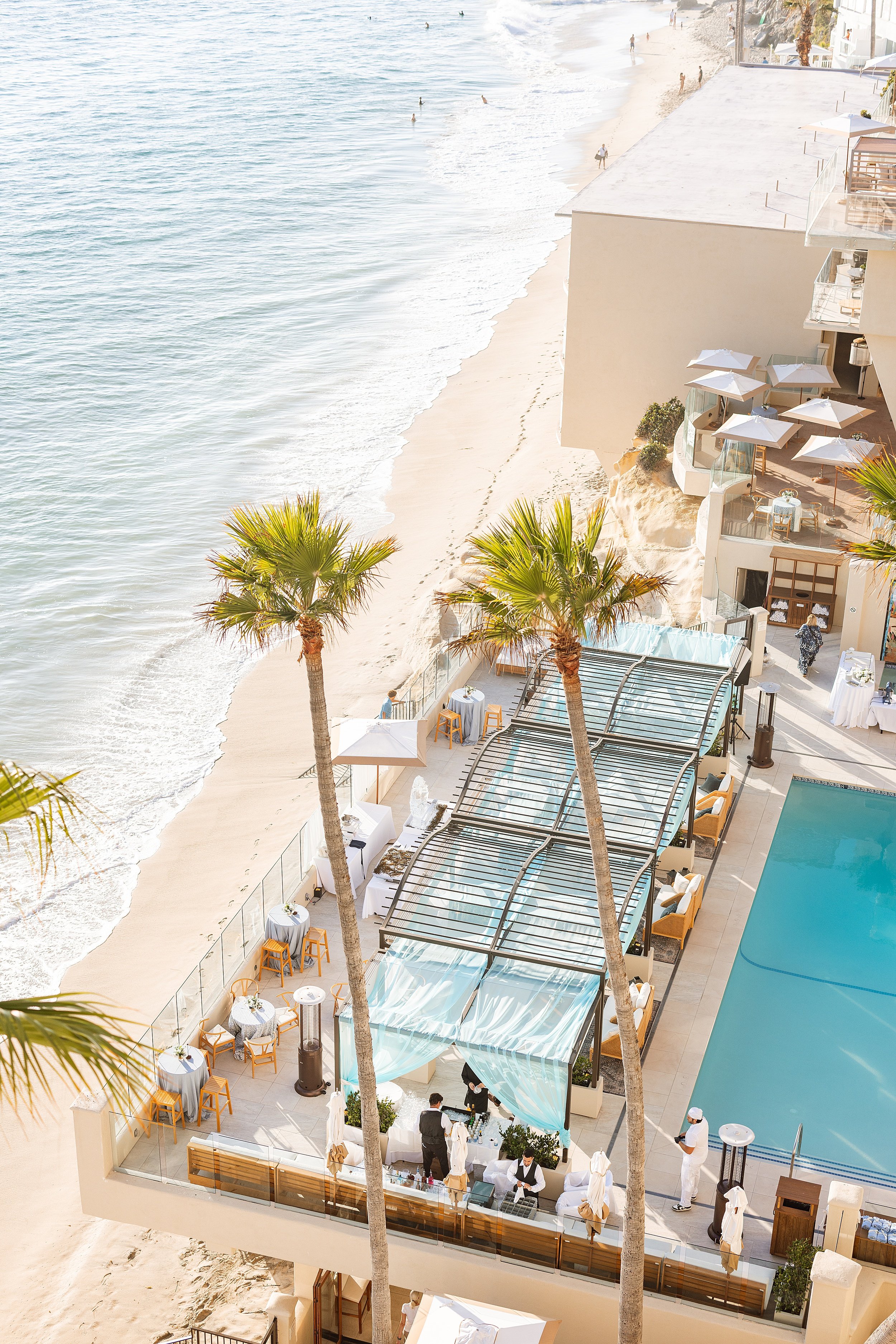 Aerial view of a beachfront hotel with a pool, outdoor seating, and palm trees, overlooking the sandy shore and ocean waves. Photo by Summerside Creative A Hotel Social Media & Creative Agency Serving Hospitality Brands