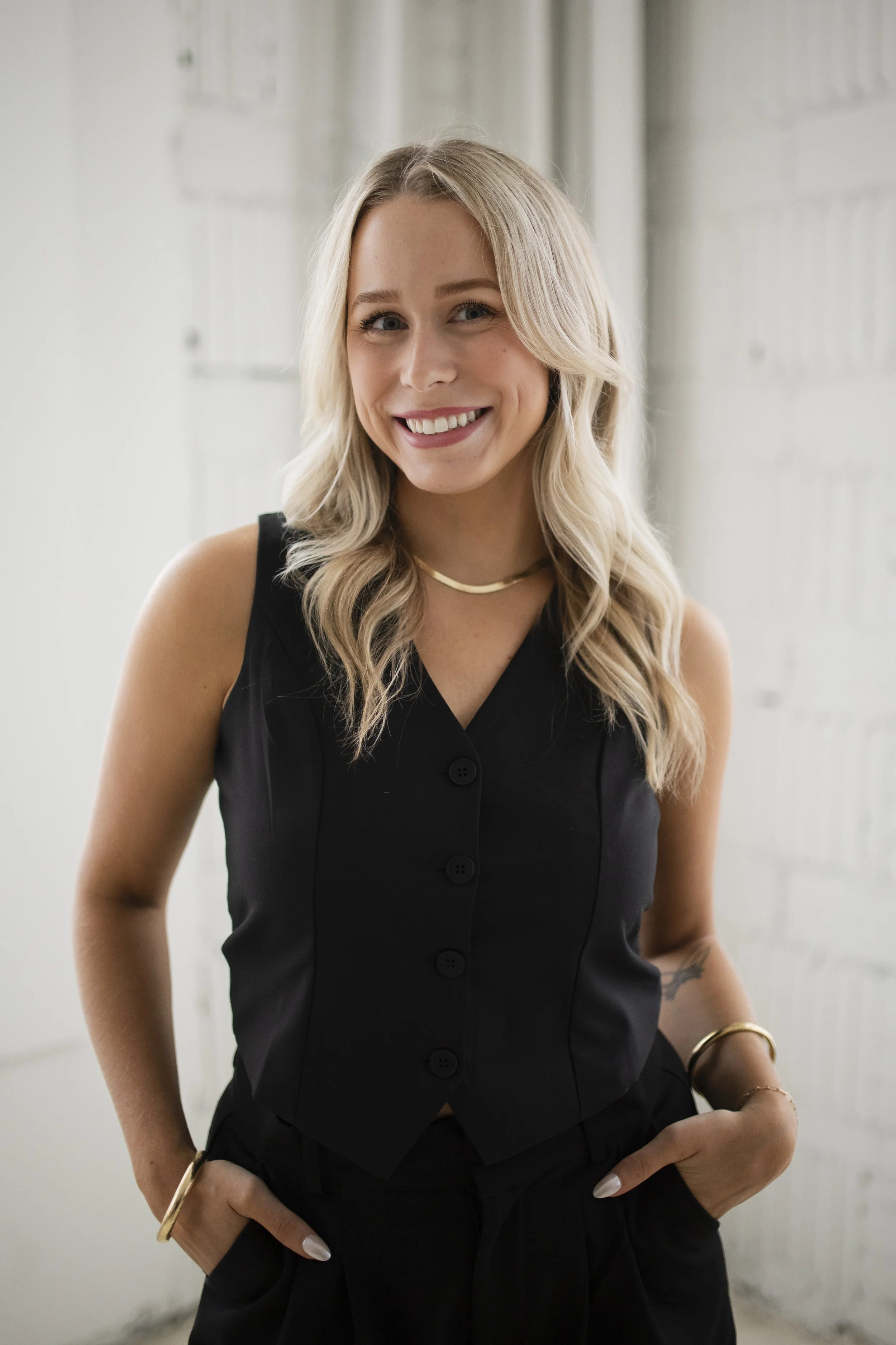 A smiling woman with blonde hair, wearing a black sleeveless buttoned top, gold jewelry, and standing in front of a light-colored brick wall.