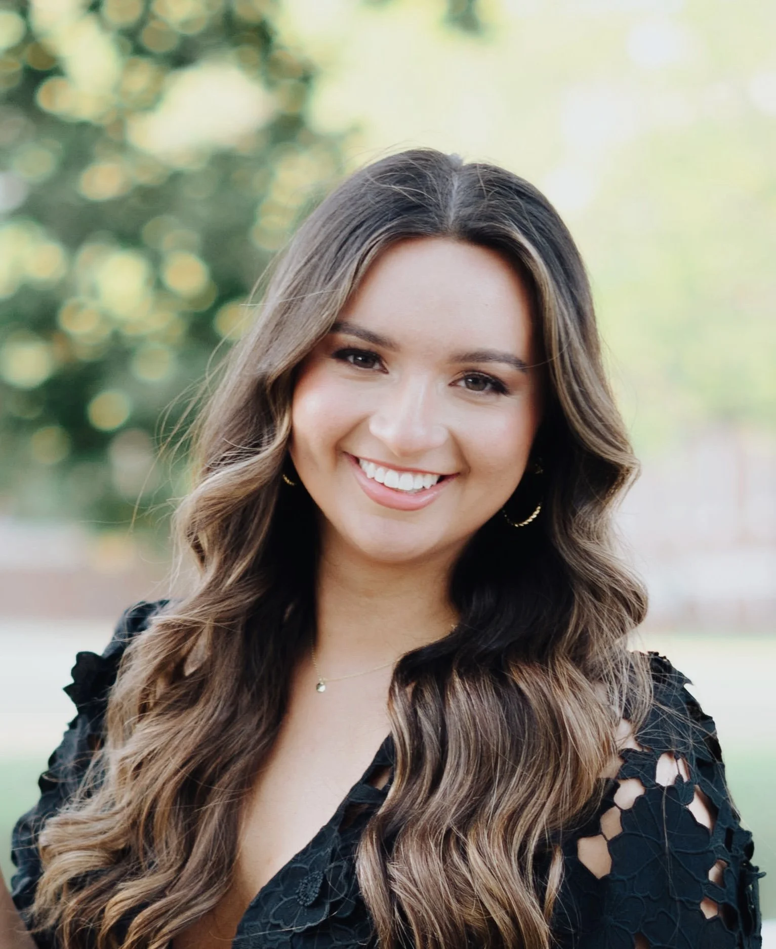 Smiling woman with long wavy brown hair outdoors, wearing a black lace top and gold jewelry.