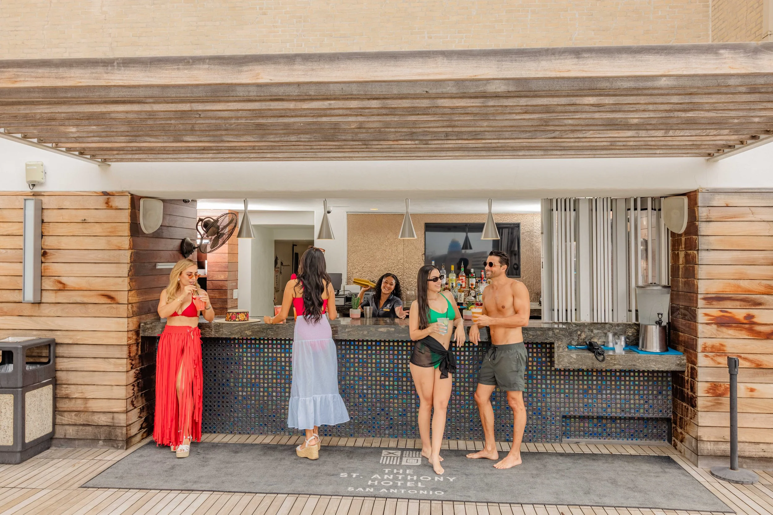Group of people at a poolside bar, two women and a man holding drinks, with two women behind the bar serving drinks, at The St. Anthony Hotel in San Antonio. Photo by Summerside Creative A Hotel Social Media & Creative Agency Serving Hospitality Bran