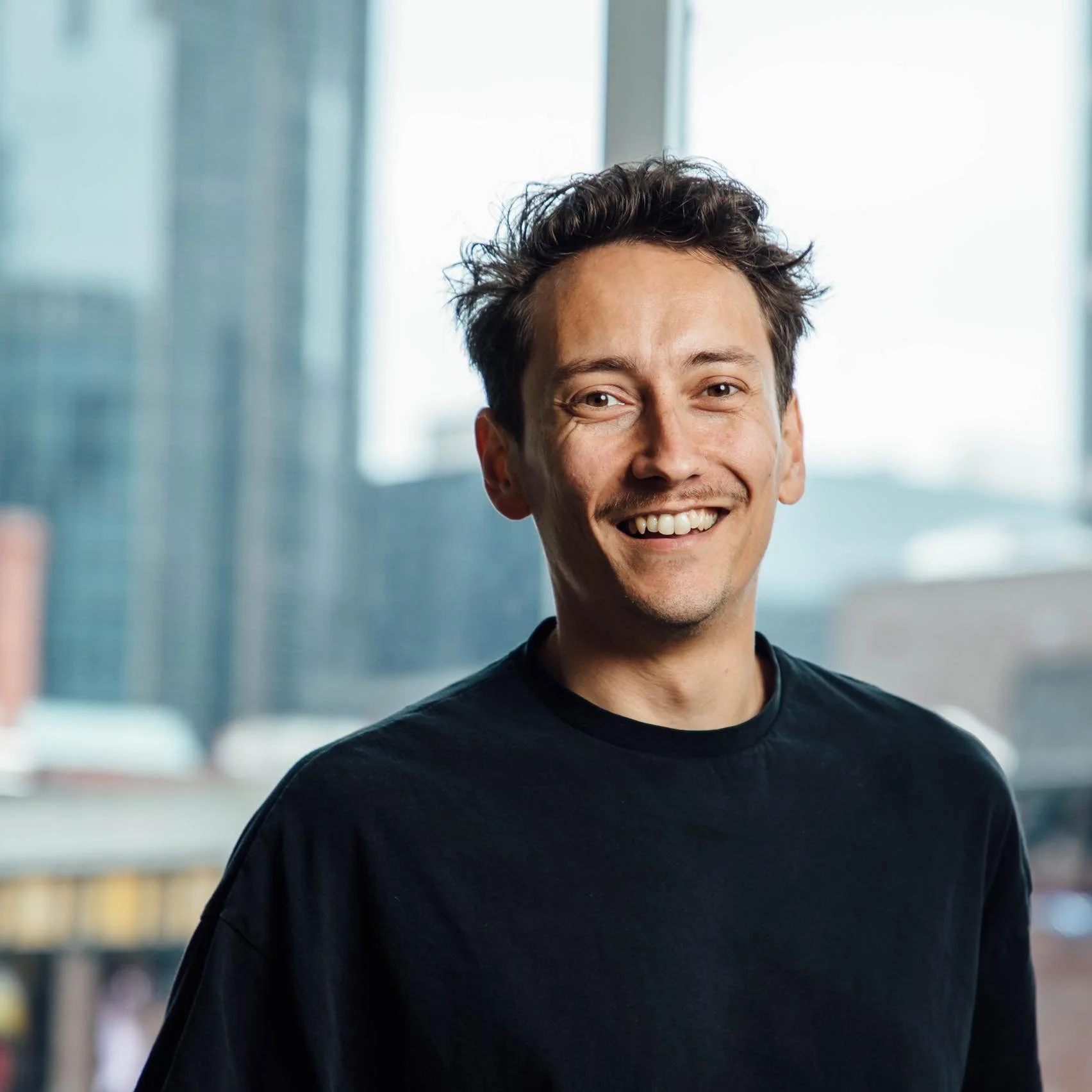 Smiling man with tousled hair wearing a black shirt, standing in front of large windows with city buildings outside.