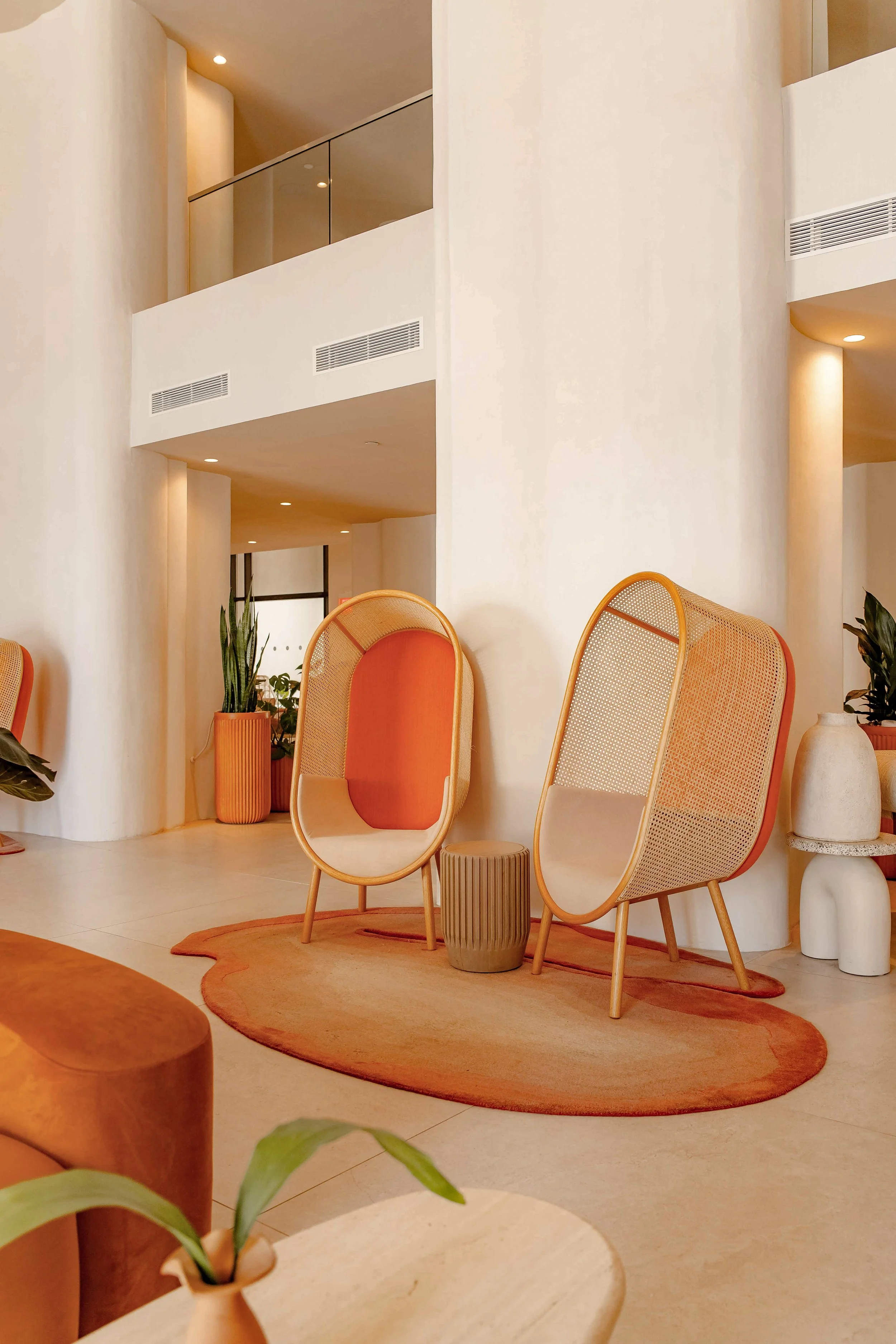 Modern hotel lobby with two unique orange and beige chairs, a small green side table, orange rug, potted plants, and white walls with rounded corners. Photo by Summerside Creative A Hotel Social Media & Creative Agency Serving Hospitality Brands