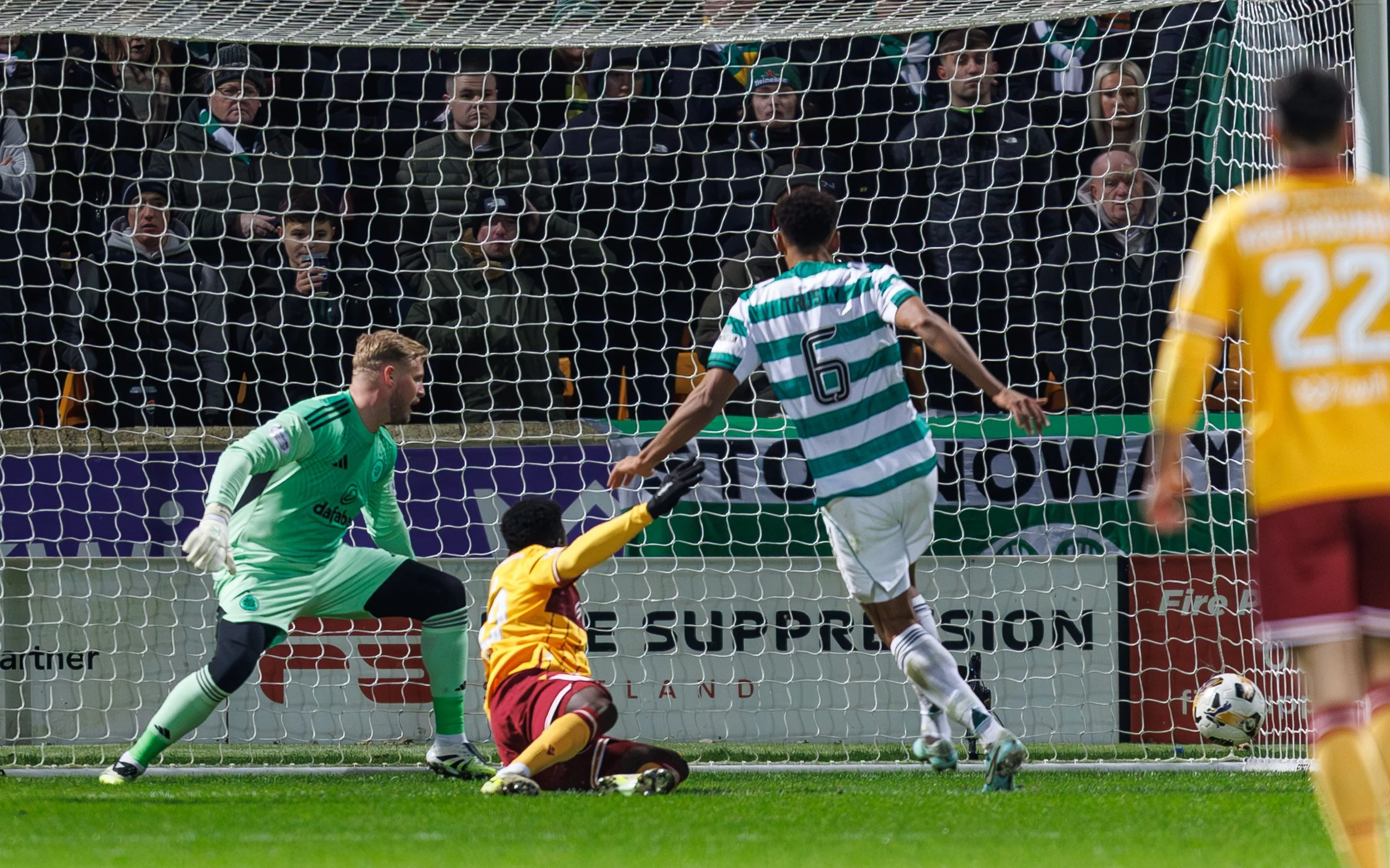 Soccer game action showing a player in a green and white striped jersey kicking the ball towards the goal as a goalie in a green uniform and a player in yellow and maroon uniforms attempt to defend. A crowd watches behind the goal.