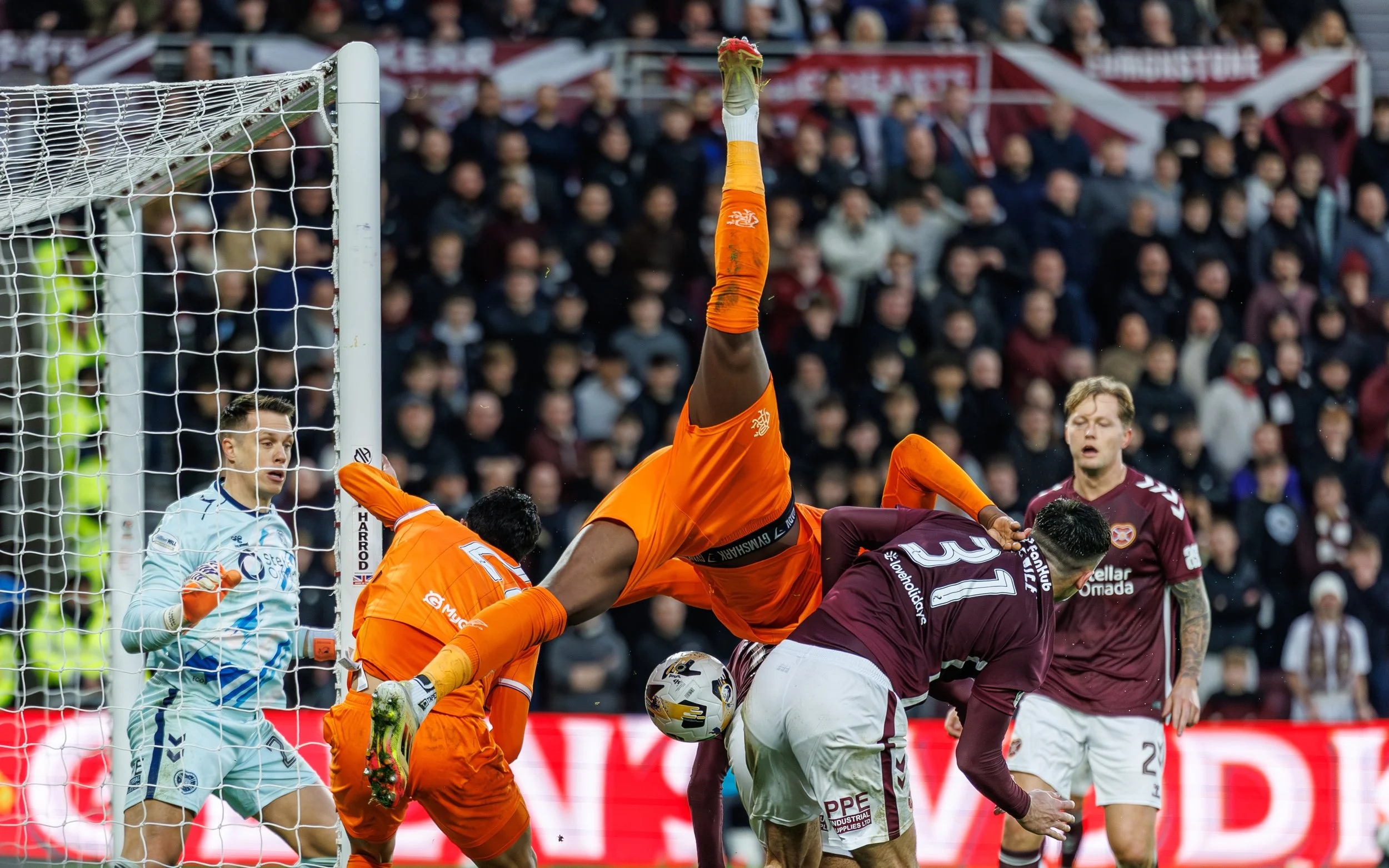 Soccer goalkeeper in teal uniform and gloves catching or blocking a ball near goal during a match, with players in maroon jerseys around him and a crowd watching in the background.