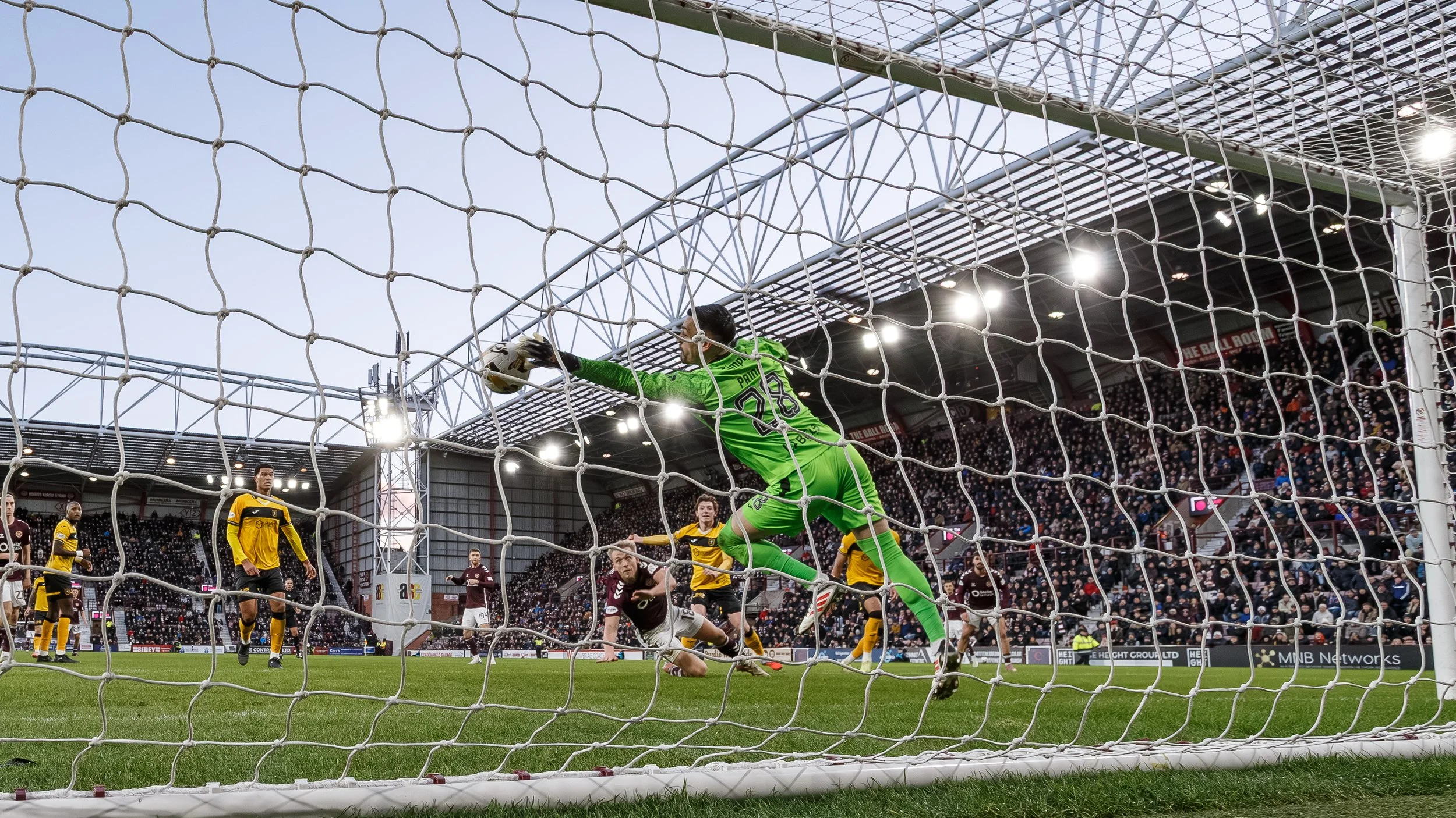 A soccer match showing a goalkeeper in a green uniform diving to catch the ball while players in maroon and yellow uniforms are nearby on the field. The stadium is full of spectators.