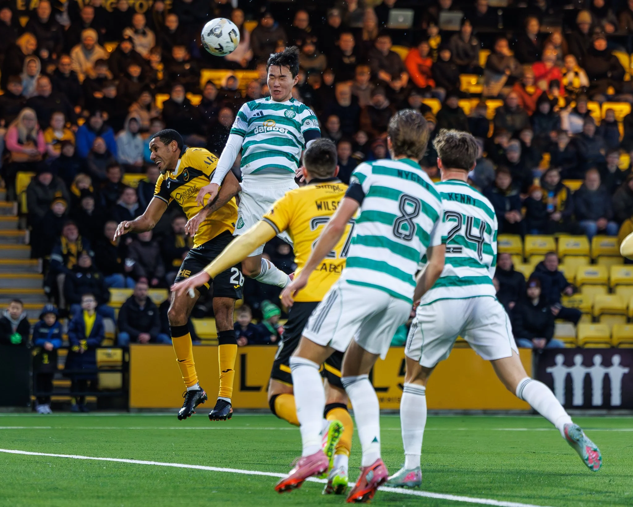 Soccer players in yellow and black uniforms and green and white uniforms are mid-play contesting for the ball during a match. Several players jump for the ball, with an audience watching from the stands.