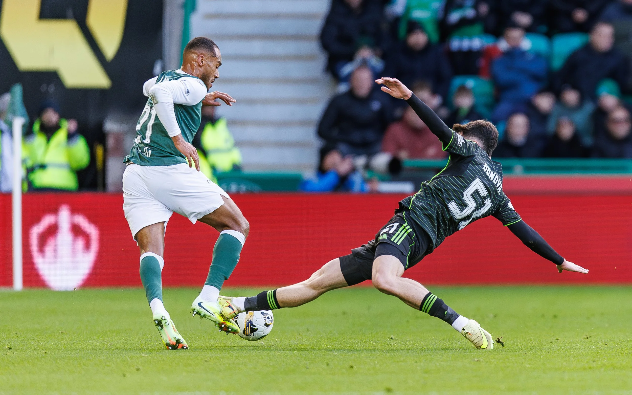 A soccer player in a green jersey and white shorts attempts to kick a ball while a goalkeeper in a black jersey and black shorts dives to block the shot during a match.