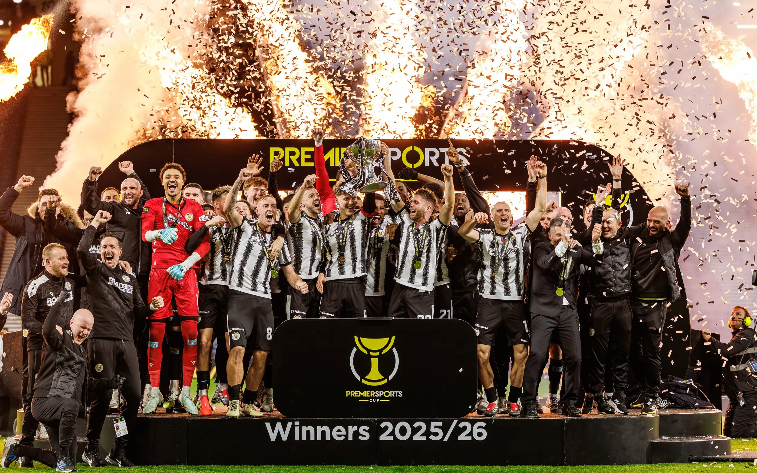 A soccer team celebrating on a podium with a trophy after winning the Premier Sports Cup in 2025/26, with fireworks and confetti in the background.