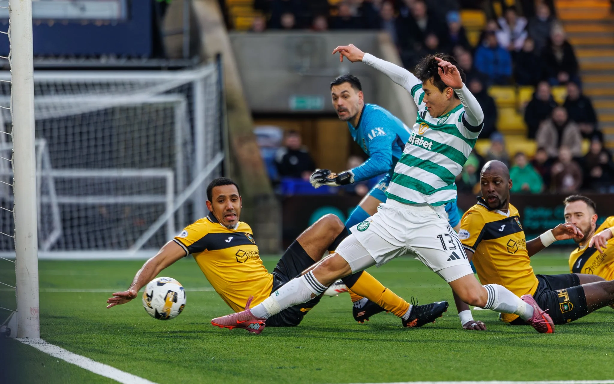 Soccer players near the goal, with one player in green and white stripes and two players in yellow and black, all on the ground, competing for the ball as a goalkeeper in blue looks on.
