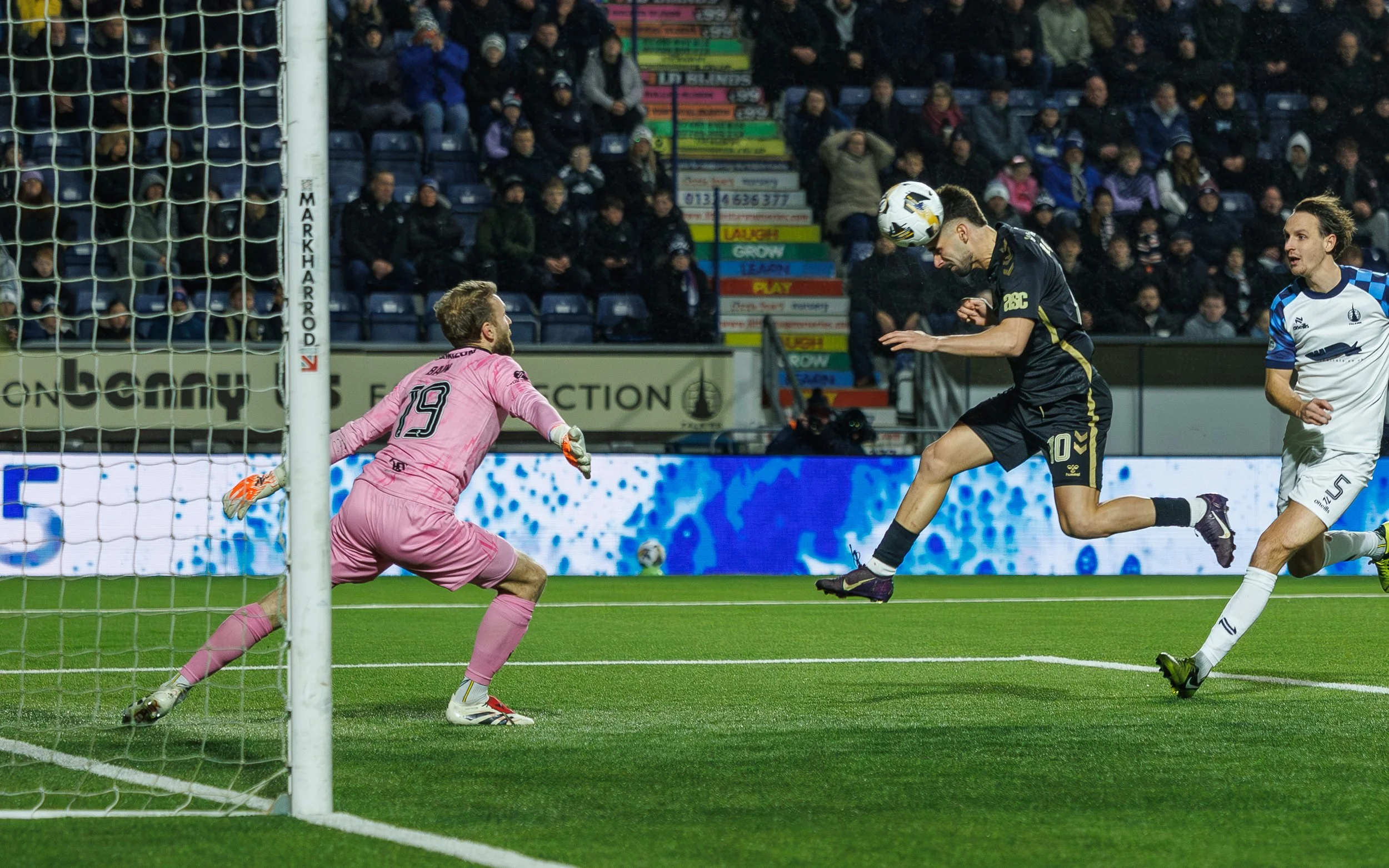Soccer match scene showing a forward in black jersey heading the ball towards the goal, with a goalkeeper in pink preparing to block, and a defender in white nearby, crowd watching in the stadium.