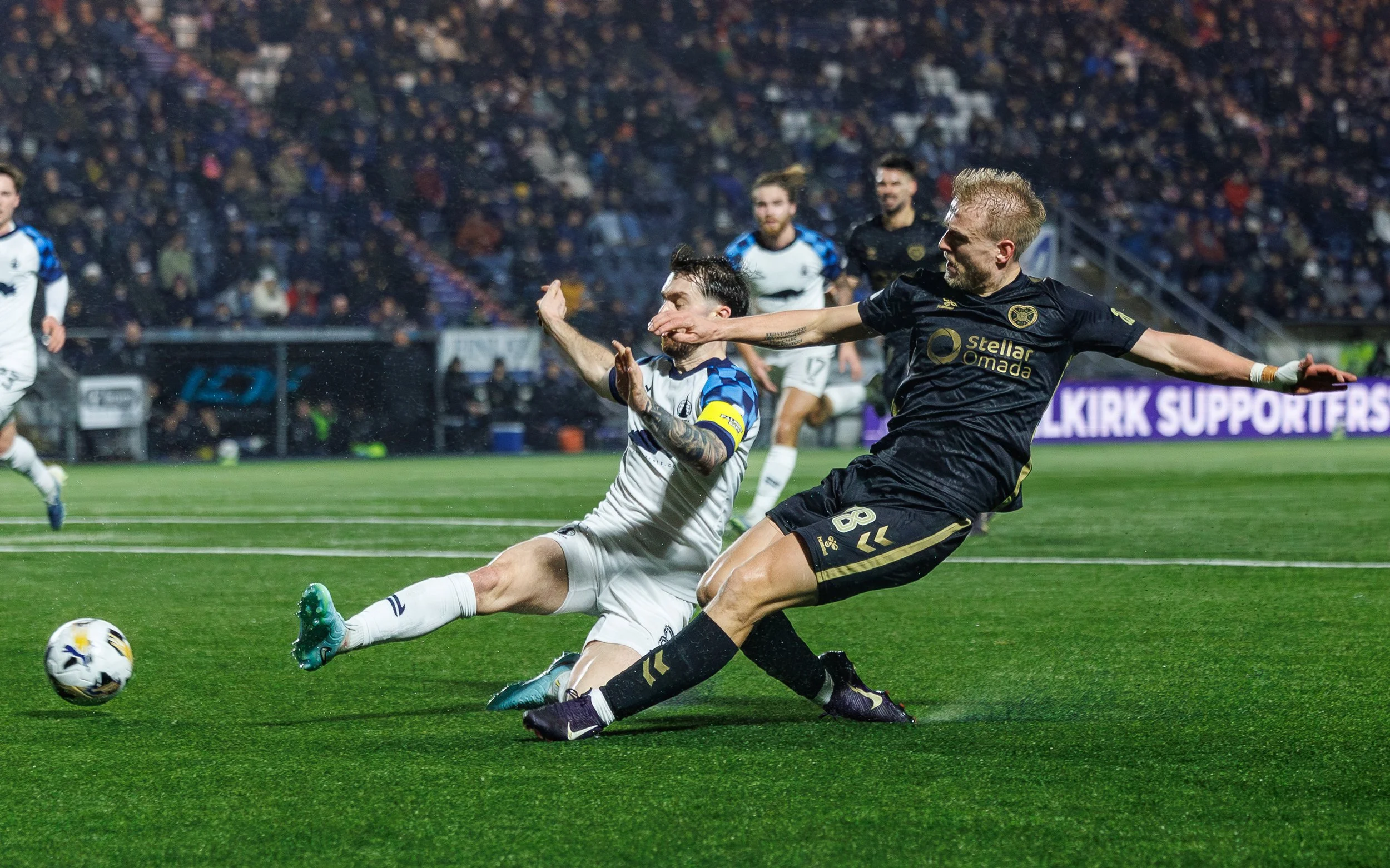 Two soccer players in a duel for the ball on a wet field during a night game, with other players and fans in the background.