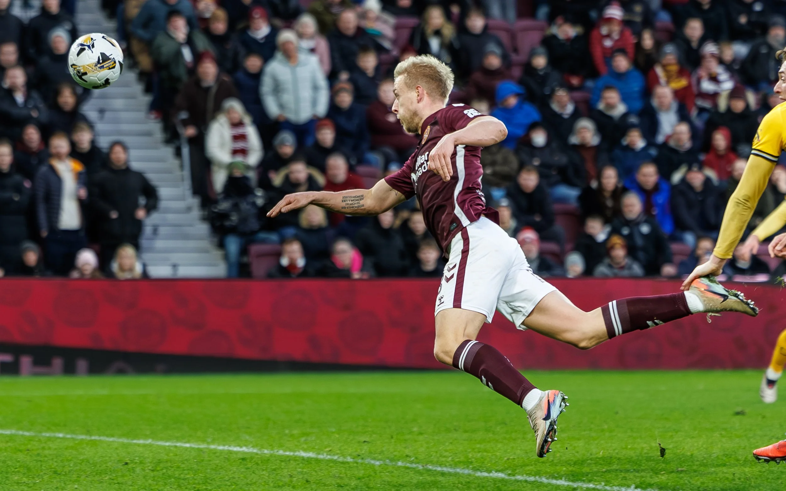A soccer player in maroon and white uniform is jumping to kick a soccer ball during a match with spectators in the background.