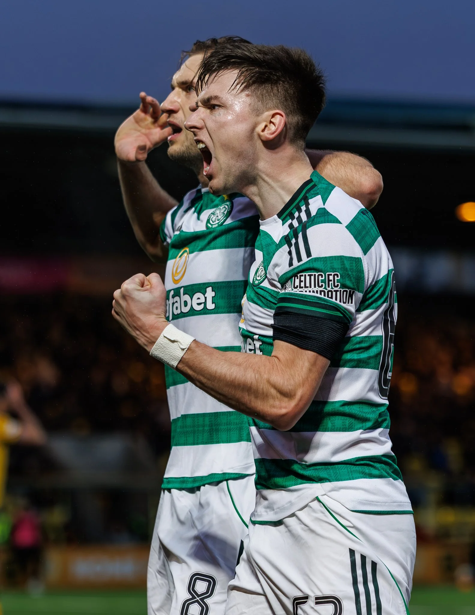 Two soccer players from Celtic FC celebrating during a match, with one player in the foreground shouting and clenching his fist.