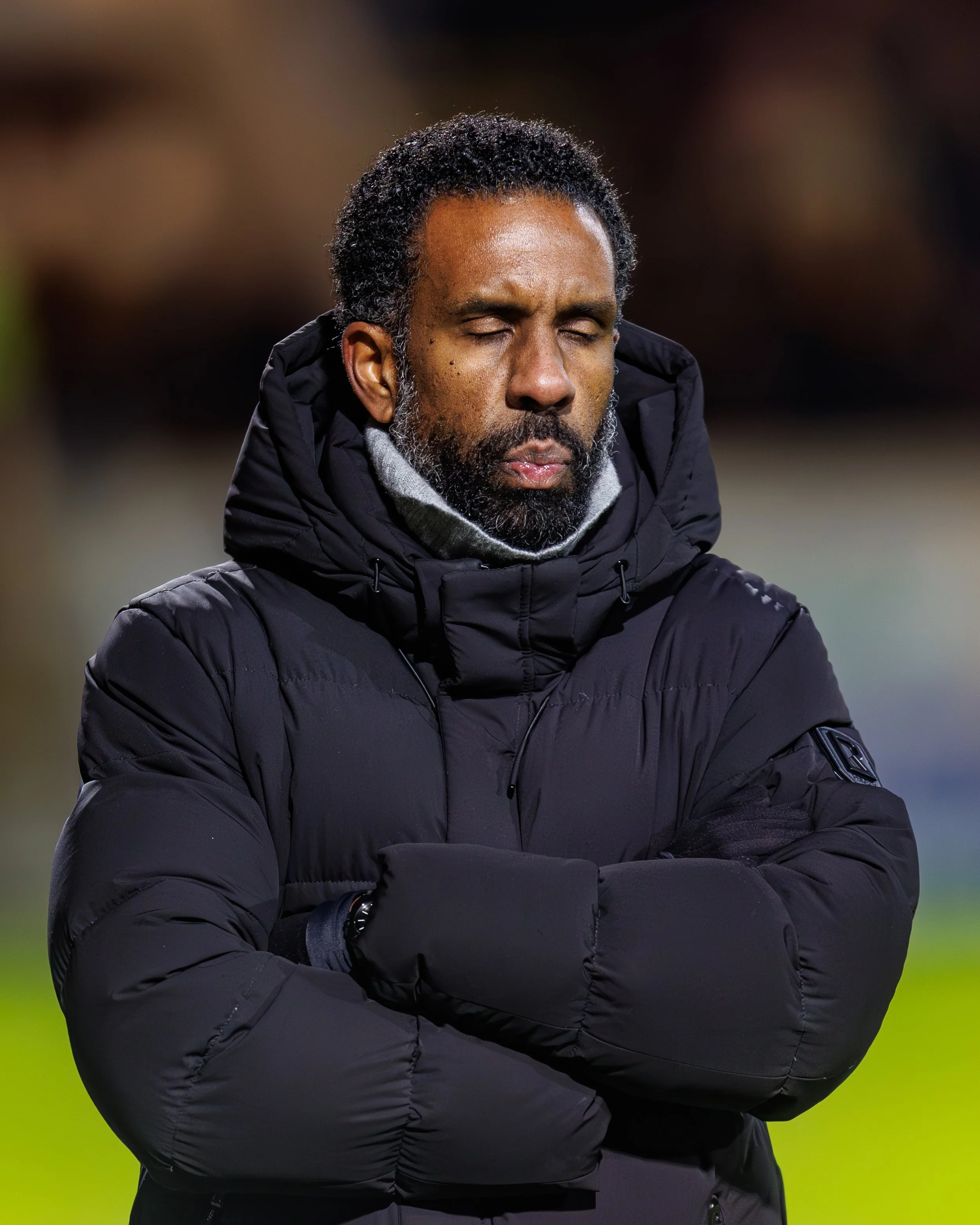 A man with dark, curly hair and a beard, dressed in a black puffer jacket with a hood, standing outdoors at night with his eyes closed and arms crossed.