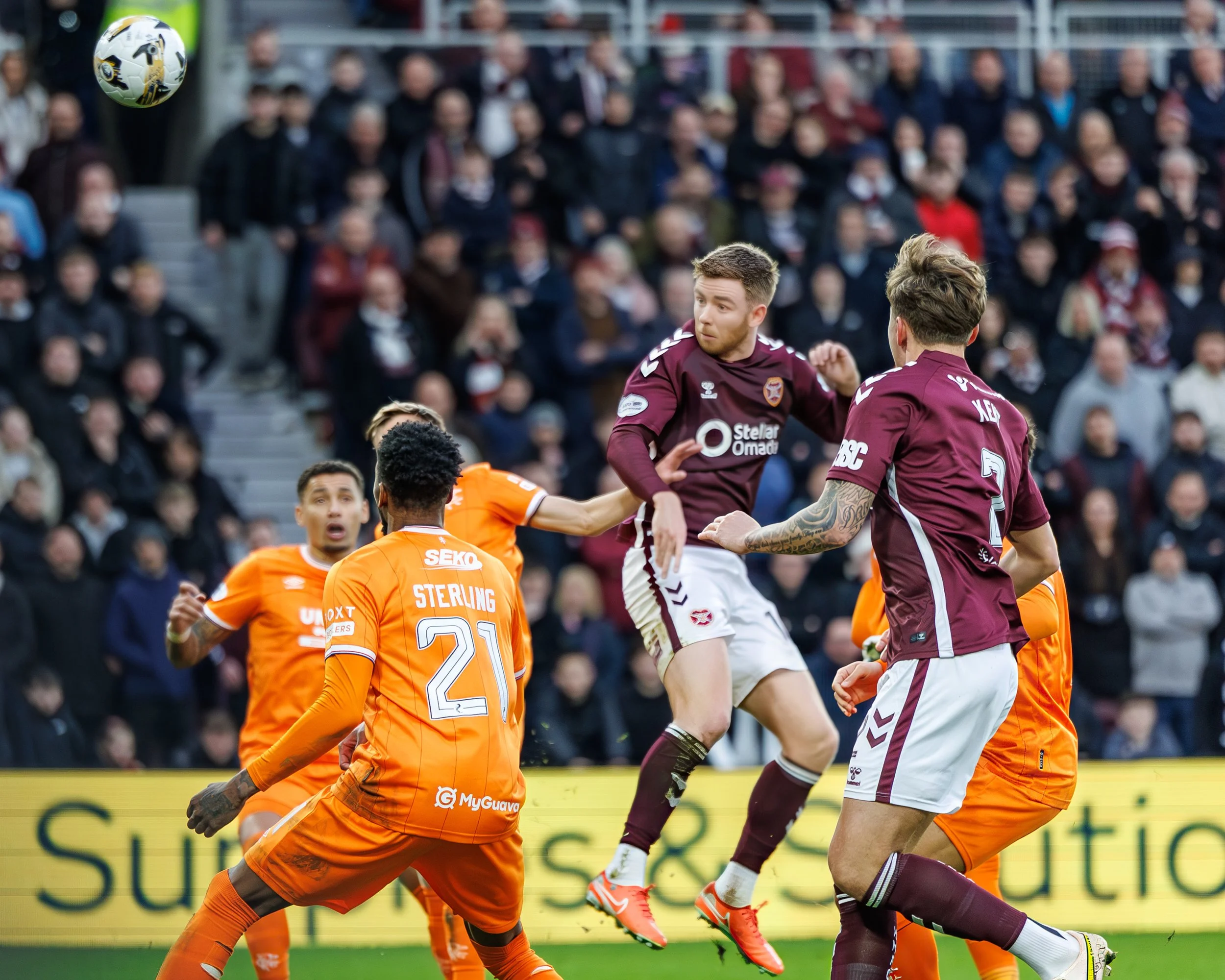A soccer match in progress with players from two teams, one in orange and the other in maroon, jumping to head the ball. The background shows a crowd of spectators watching the game.