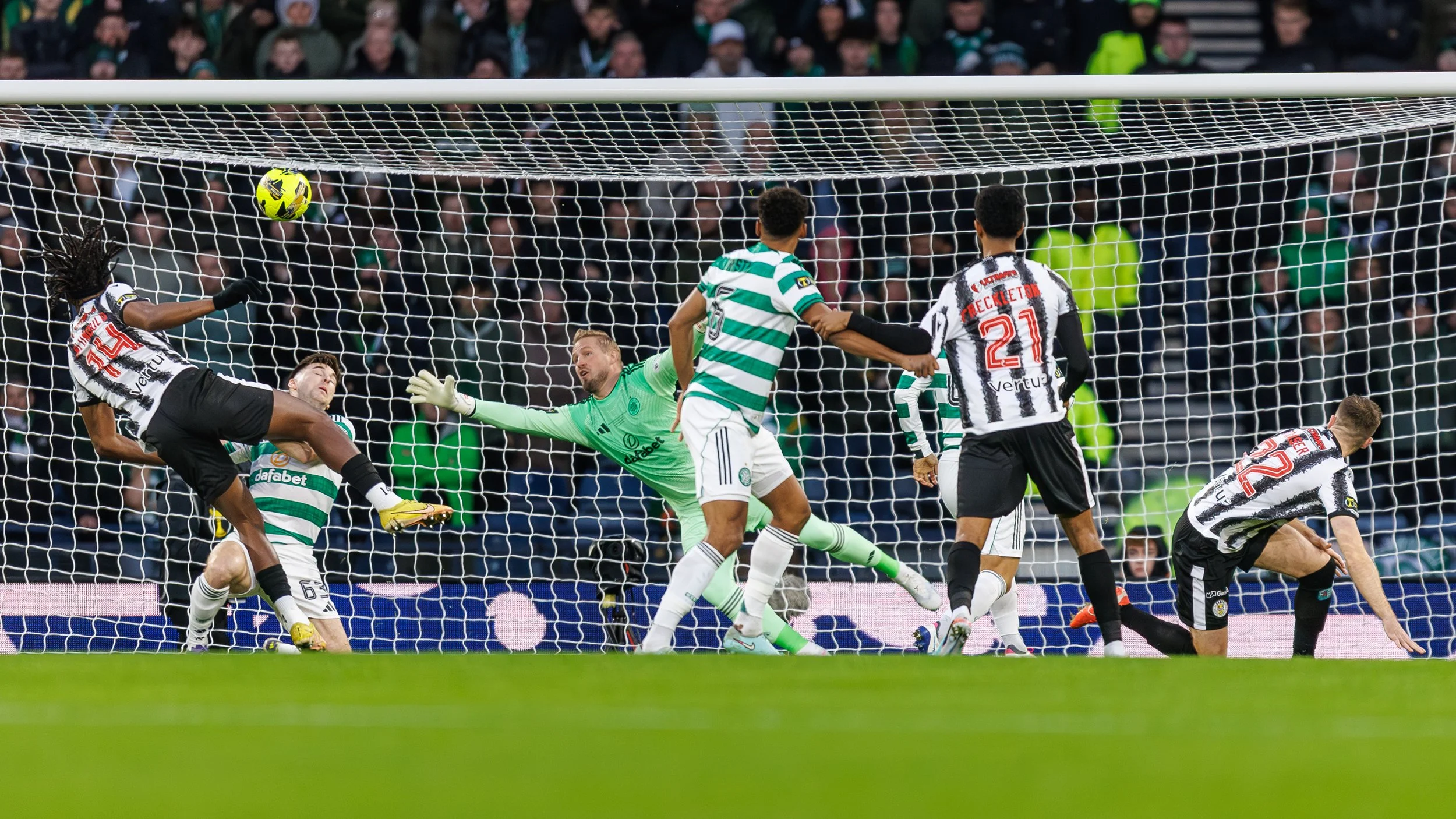 Soccer players from two teams near the goal, with one player attempting to score as the goalkeeper blocks the shot. The scene captures a moment of intense action during a match.