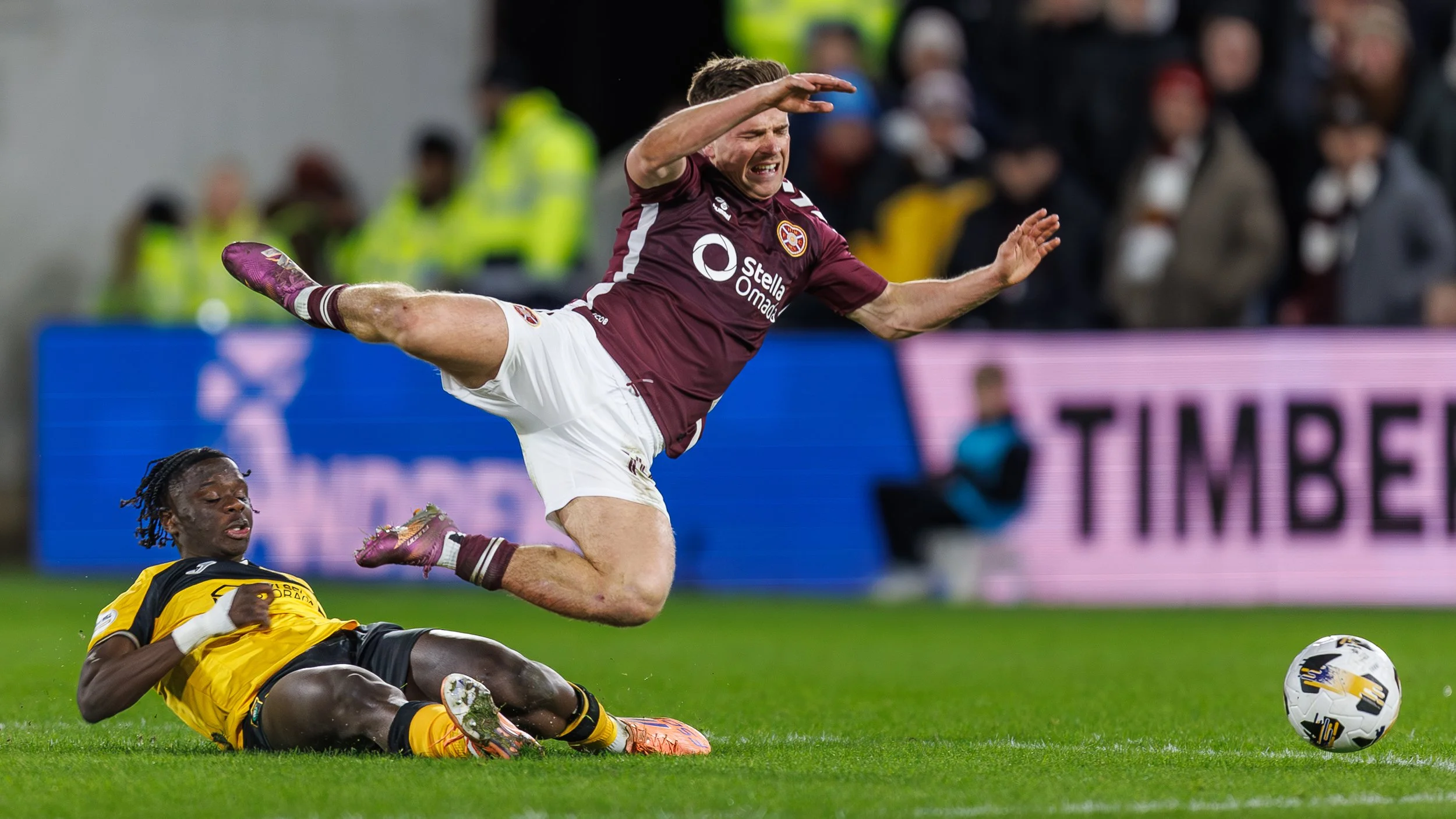 A soccer player in a maroon jersey leaps over a player in a yellow jersey who is on the ground during a match, with a soccer ball nearby on the grass field and spectators in the background.