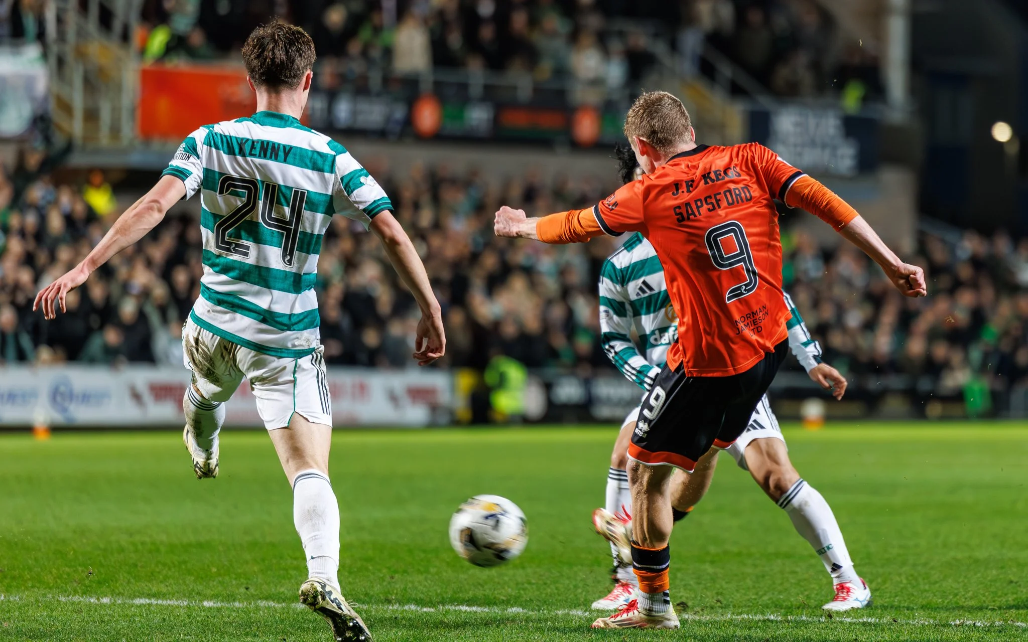 Two soccer players compete for the ball during a match, with a crowd in the background.