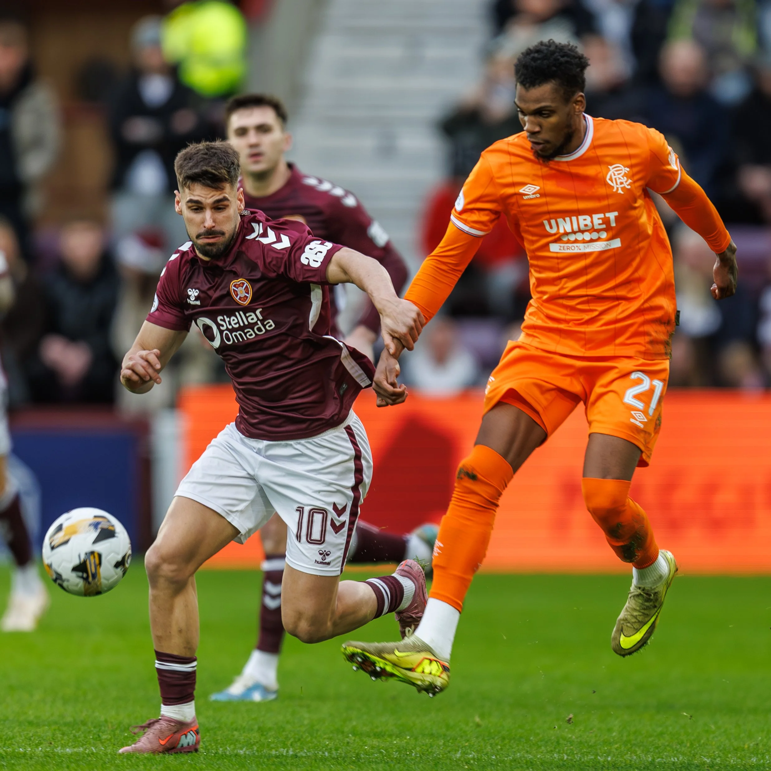 Two soccer players from opposing teams compete for the ball during a match, with one player in maroon jersey and white shorts, and the other in bright orange jersey and shorts.