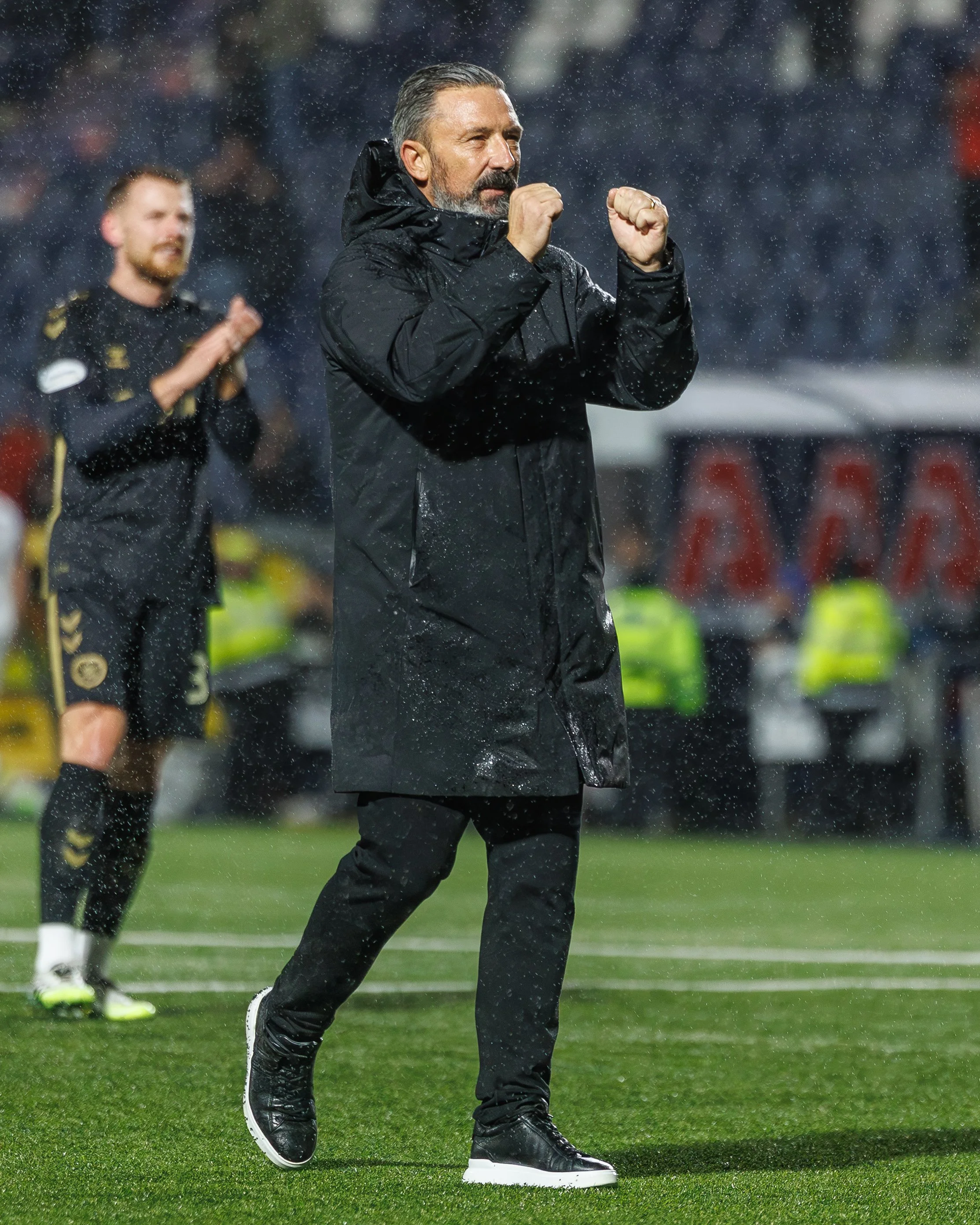 A man in a black raincoat and black shoes claps his fists in front of him on a football field during rain, with a player and security personnel in the background.