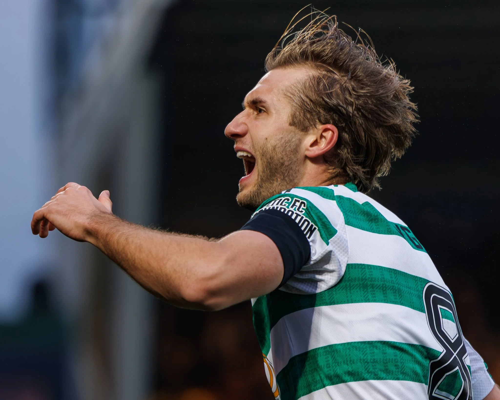A male soccer player in a green and white striped jersey celebrating with his mouth open, arms raised, and hair tousled during a match.