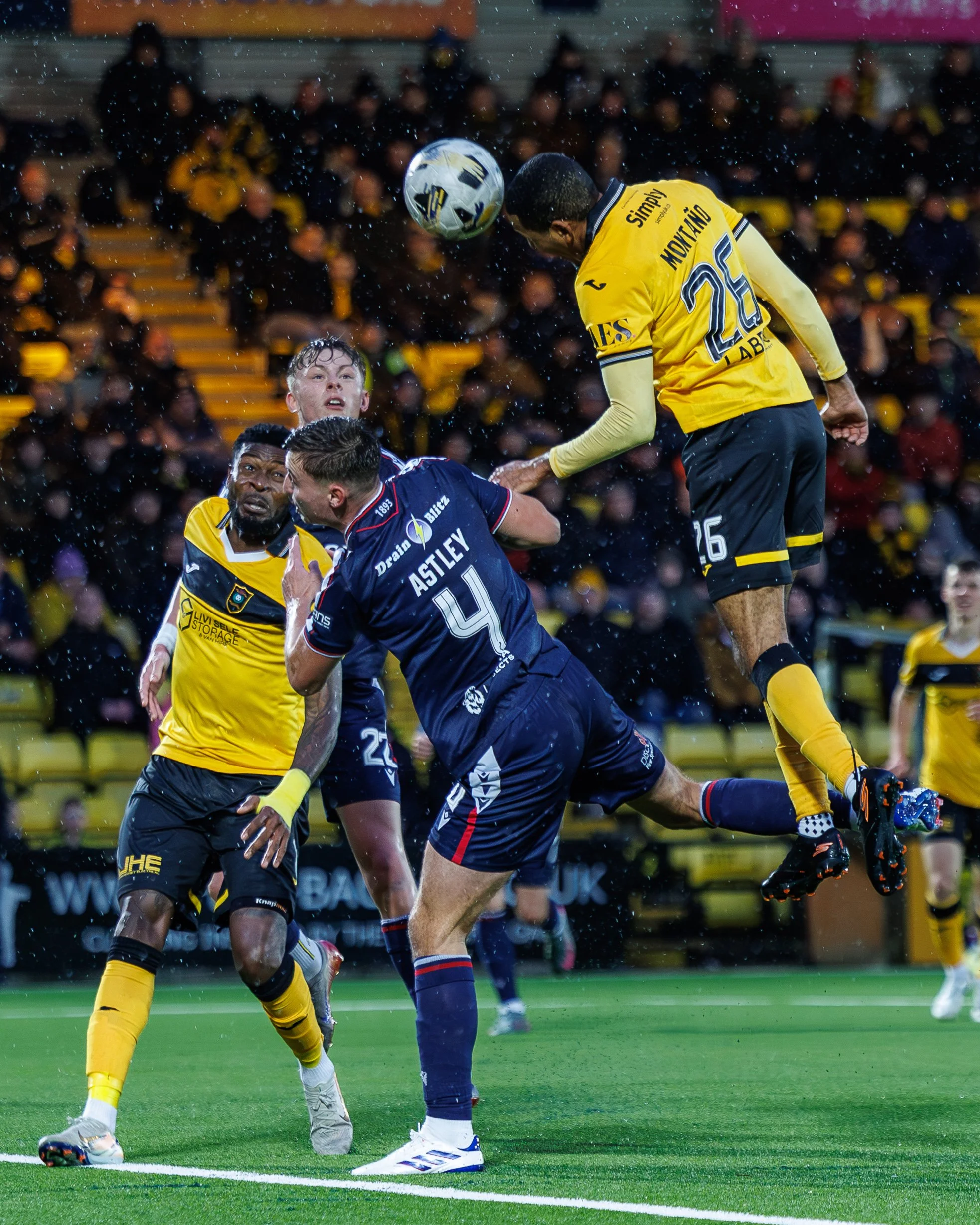 Soccer players in yellow and blue jerseys compete for the ball in a rain-soaked match, with one player jumping to head the ball while others attempt to block or kick.