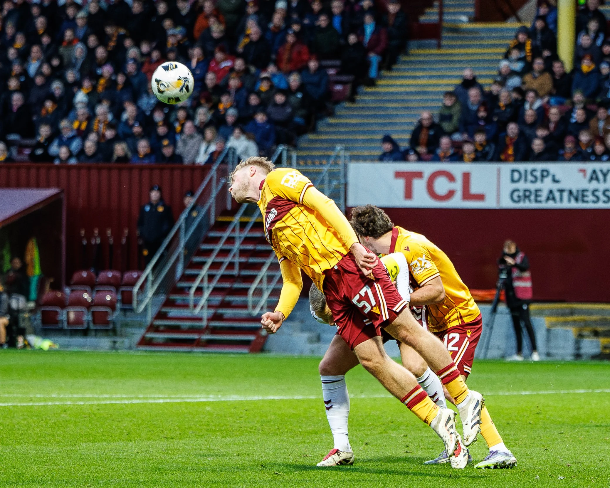 Two soccer players in yellow and red uniforms jumping to head a soccer ball during a match, with spectators in the background.