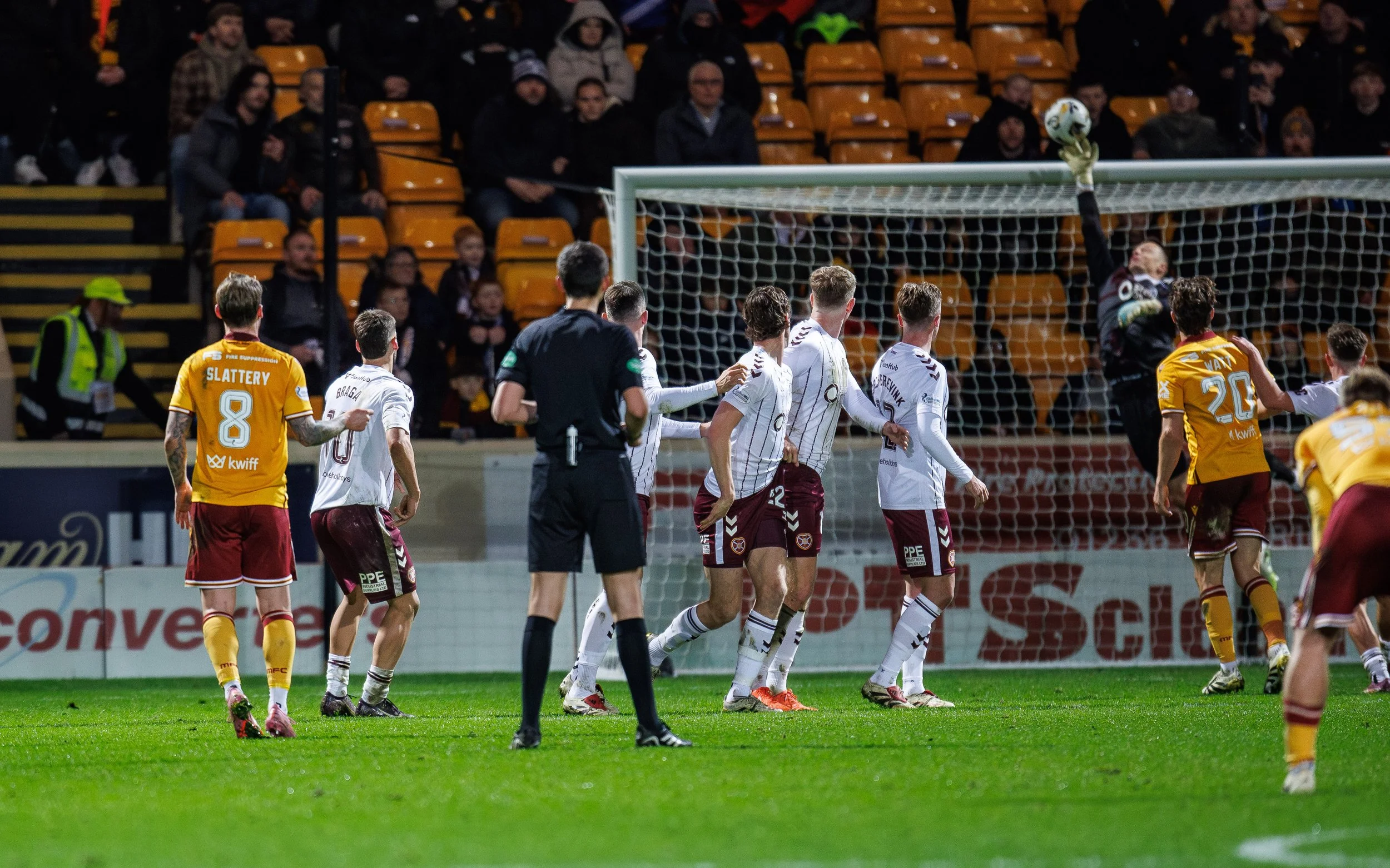 Soccer players on the field during a match, with a goalkeeper jumping to catch the ball near the goal, and spectators watching from the stands.