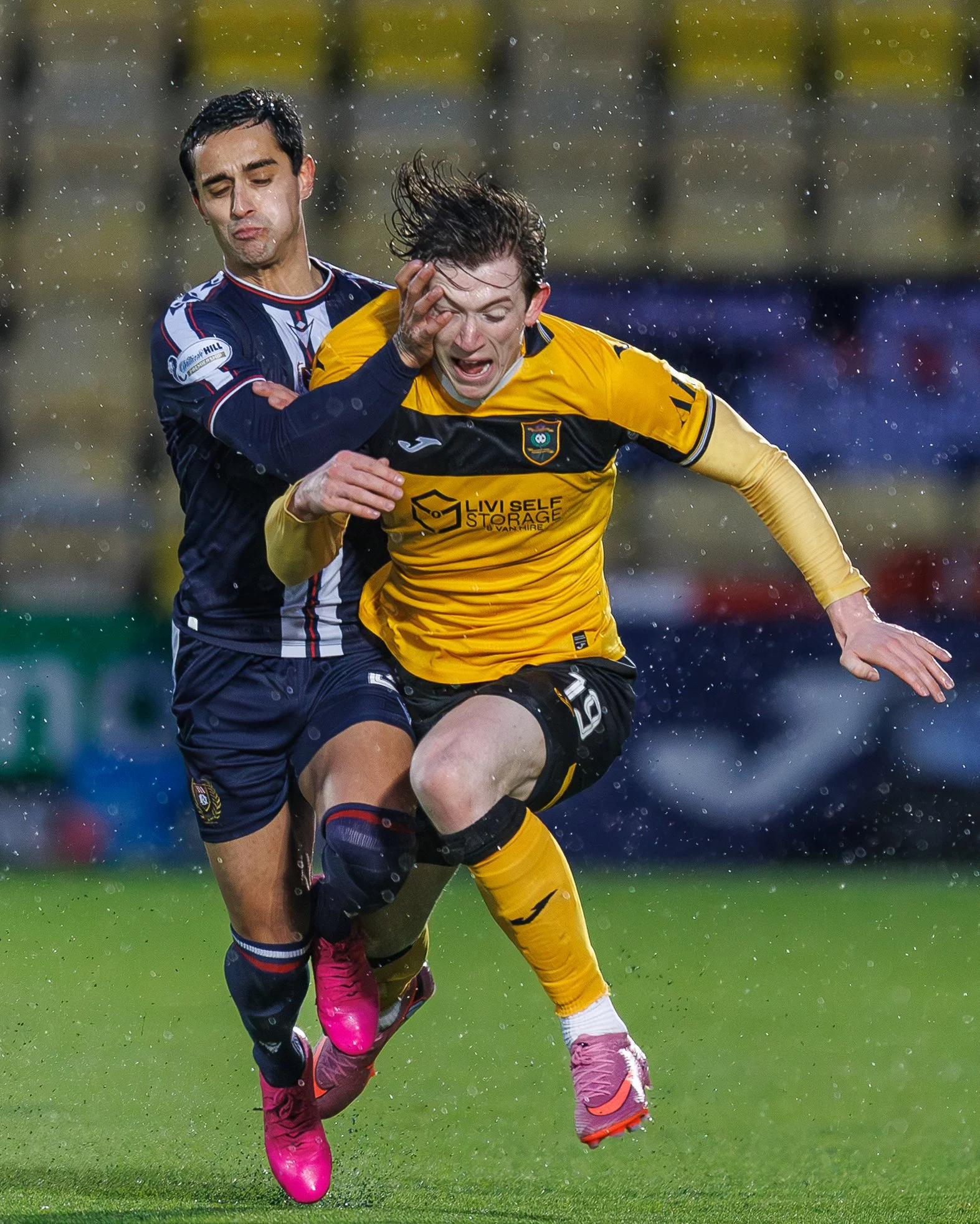 Two soccer players competing for the ball during a game in rainy weather. One player is wearing a yellow jersey, and the other is in a dark blue jersey. The yellow jersey player is running while the blue jersey player is trying to block or tackle.