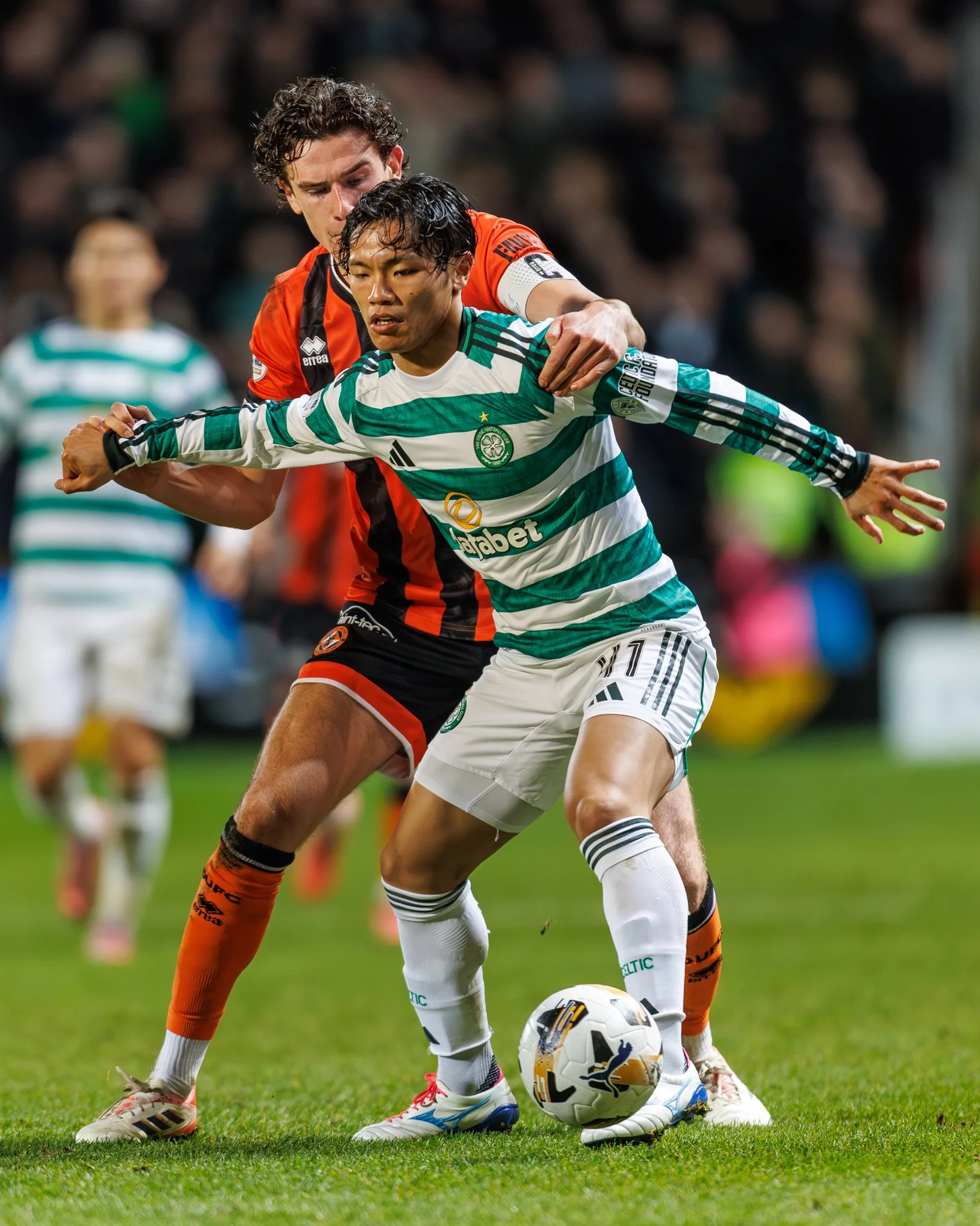 Two soccer players compete for the ball during a match, one wearing a green and white striped jersey and the other in an orange and black jersey, with a crowd in the background.