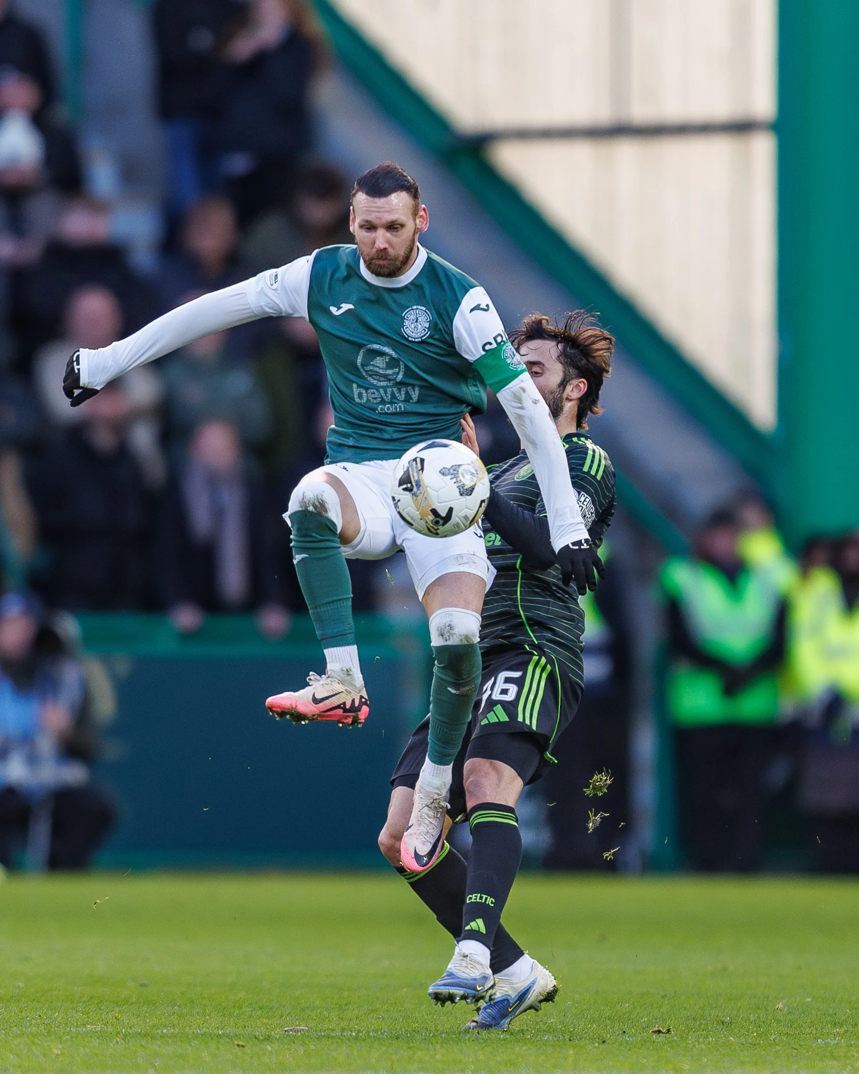 Two soccer players competing for the ball during a match; one in a green and white uniform jumping and the other in a black and green uniform attempting to block.