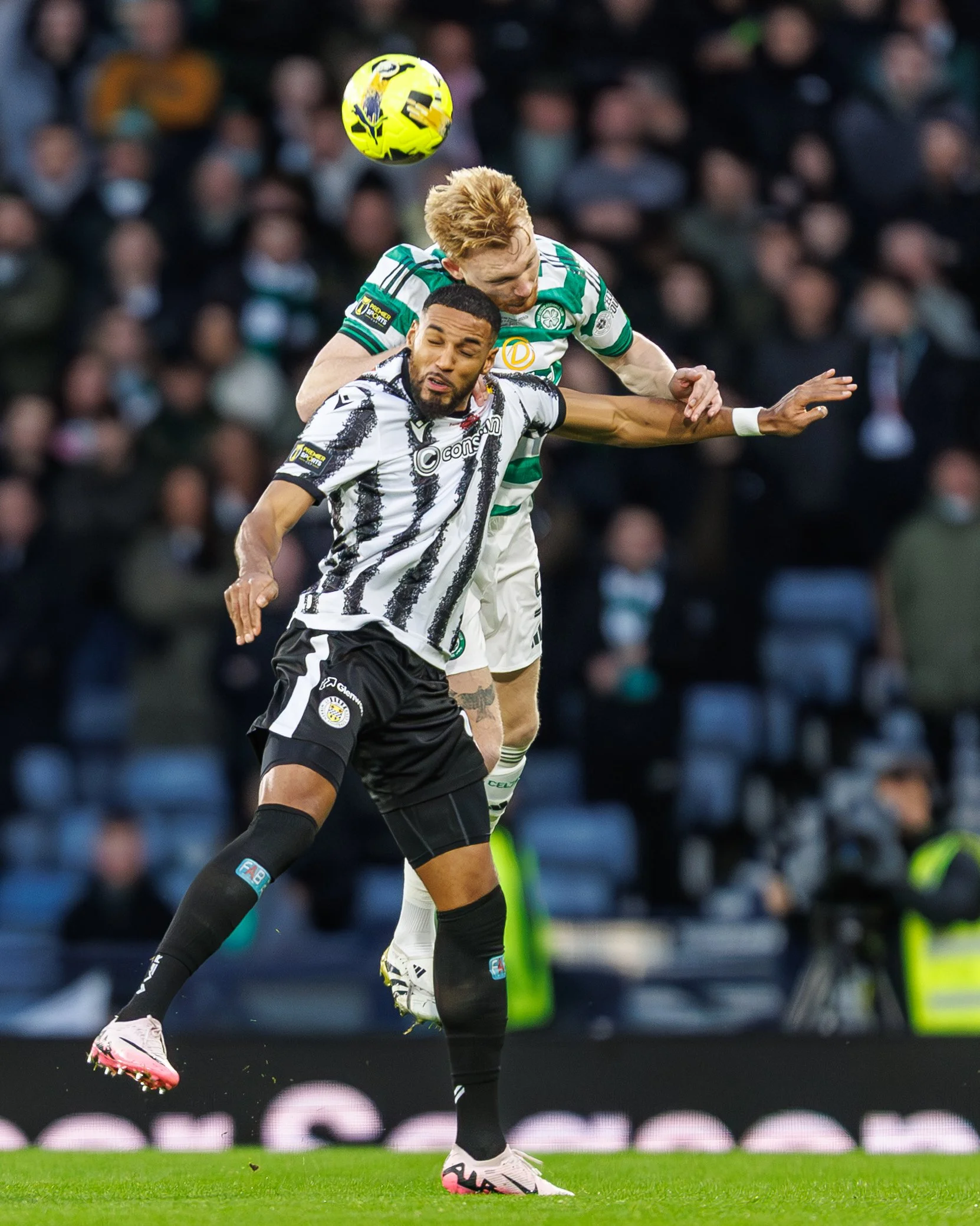 Two soccer players, one in a black and white striped jersey and the other in a green and white striped jersey, jump to head a yellow and black soccer ball during a match, with a blurred crowd in the background.