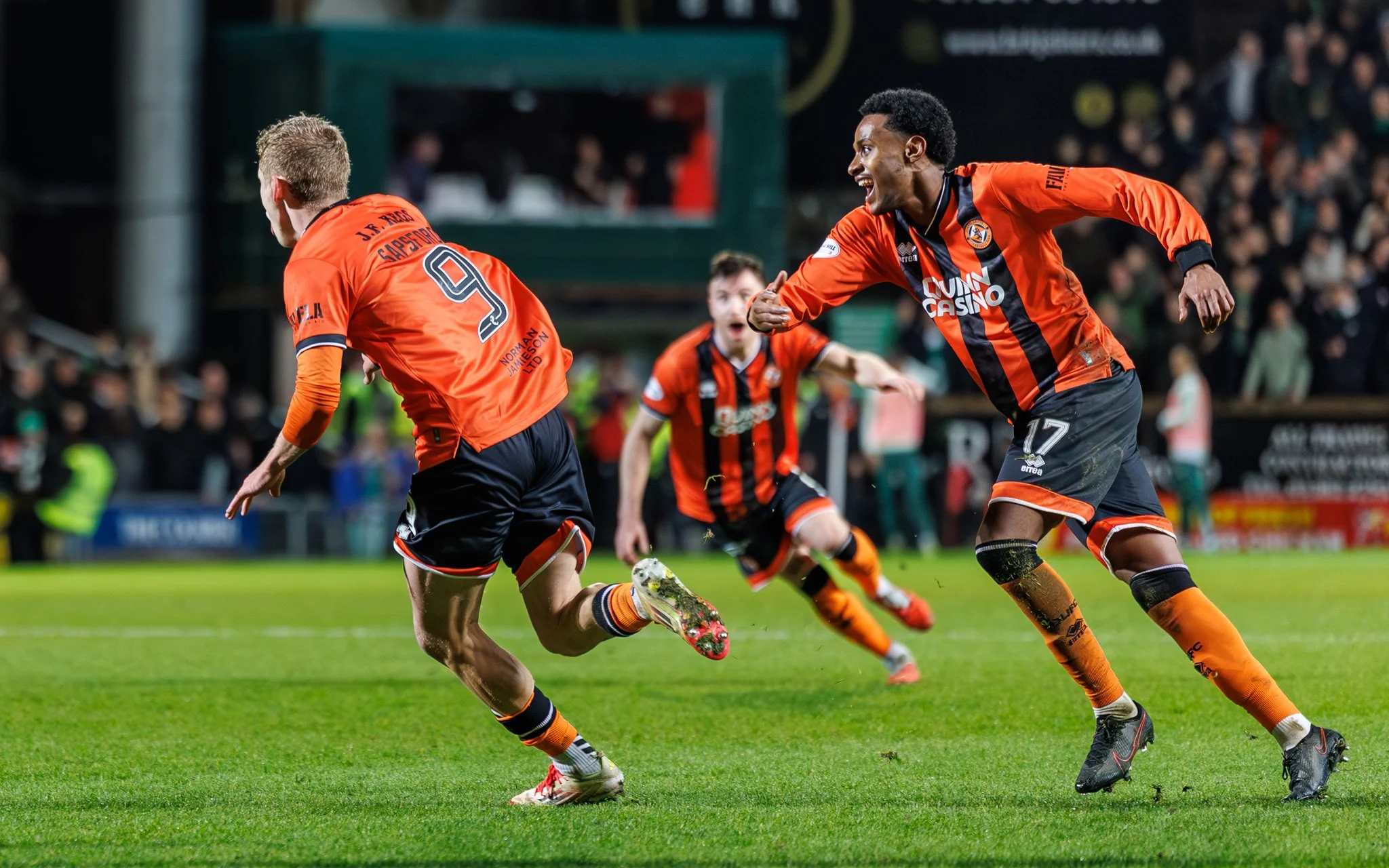 Soccer players celebrating on the field during a match, wearing orange and black uniforms, with one player running ahead and others cheering and running behind.