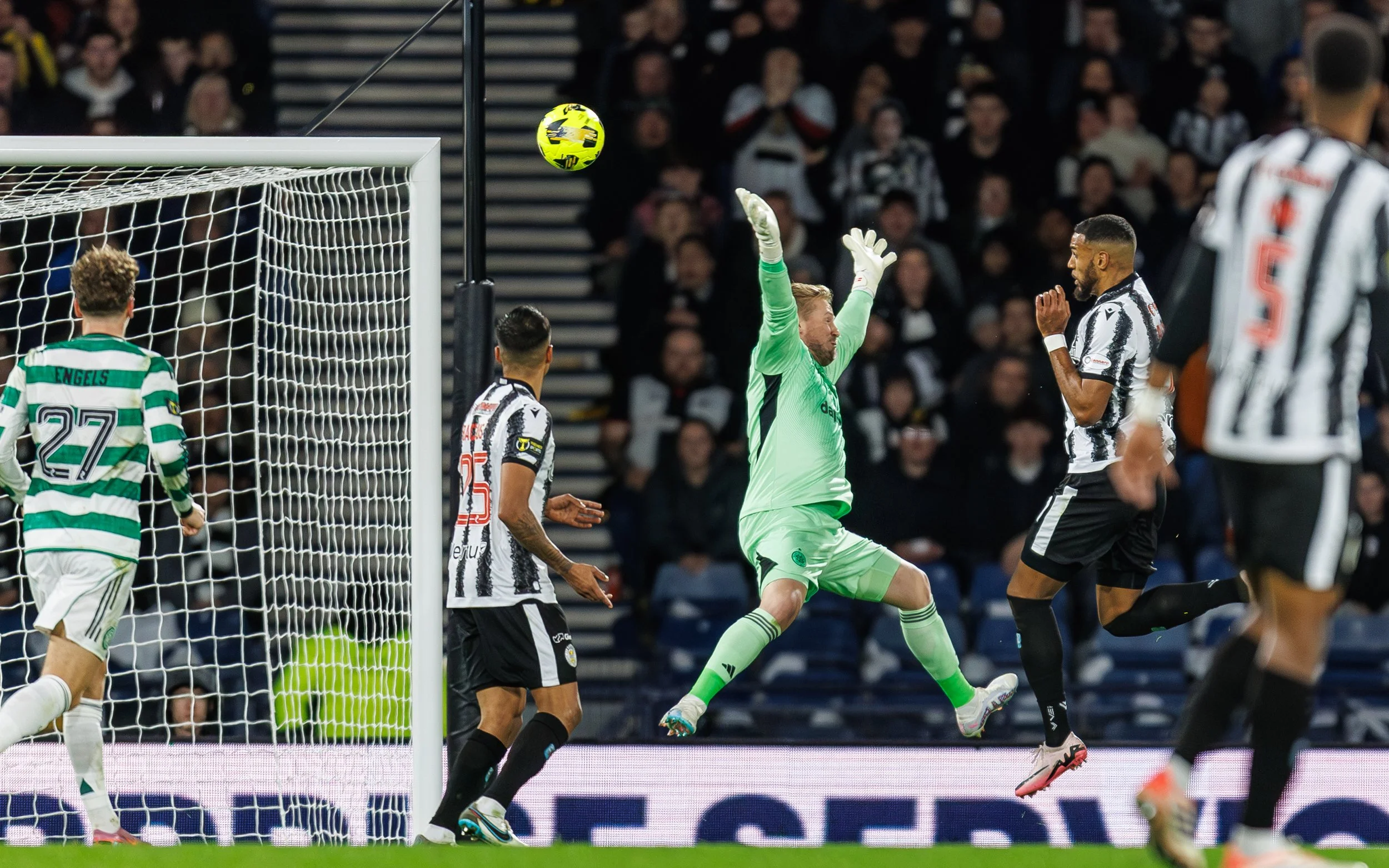 Soccer players in black and white striped jerseys compete for ball near the goal, with the goalkeeper in bright green attempting to block the shot, in a stadium filled with spectators.