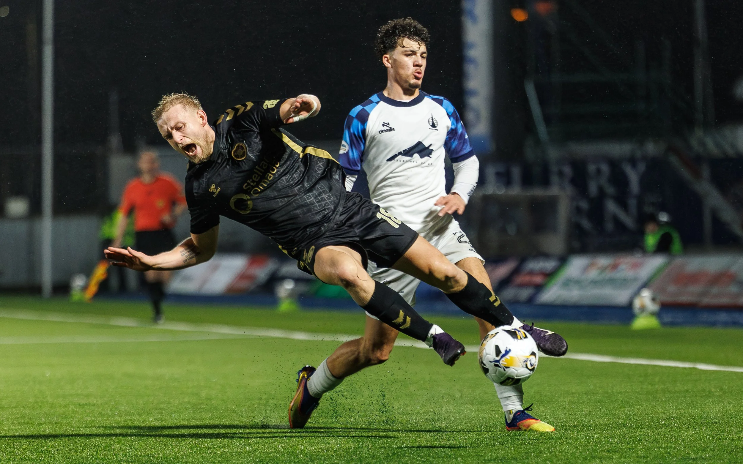 Two soccer players competing for the ball during a match. One in black uniform falling while the other in a white and blue uniform standing.