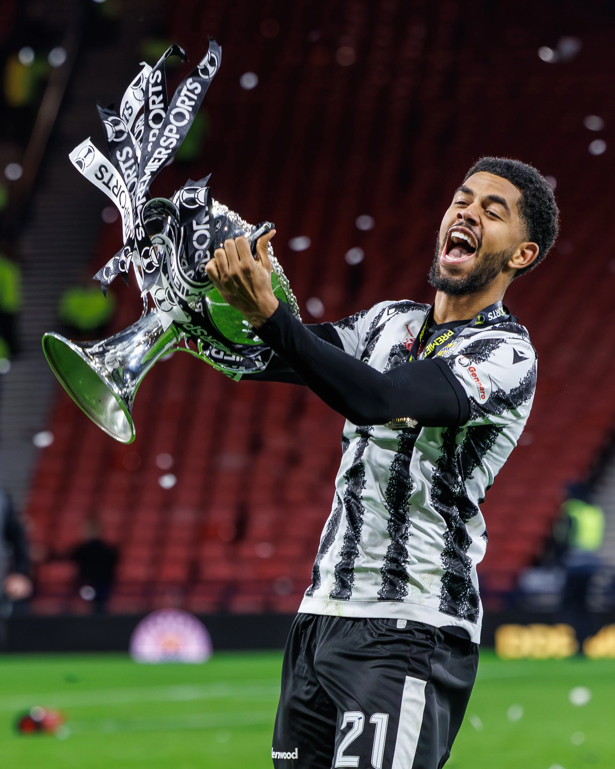 A man celebrating on a soccer field holding a trophy, with confetti falling around him, wearing a black and white jersey with the number 21.